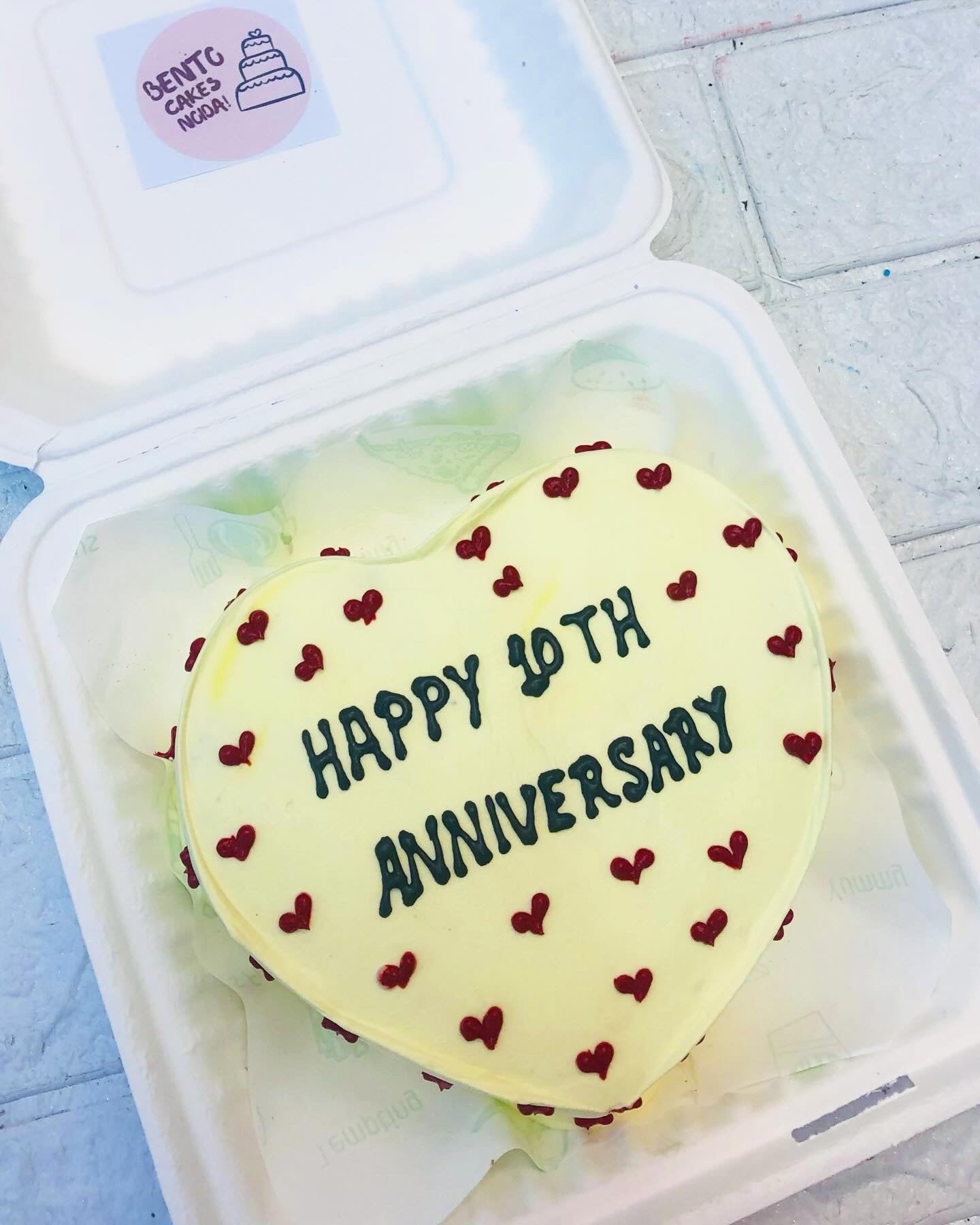 AN Offwhite heart shape acke with little red hearts design all over the cake and a text "HAPPY 10th ANNIVERSARY" wiritten with black icing on it.