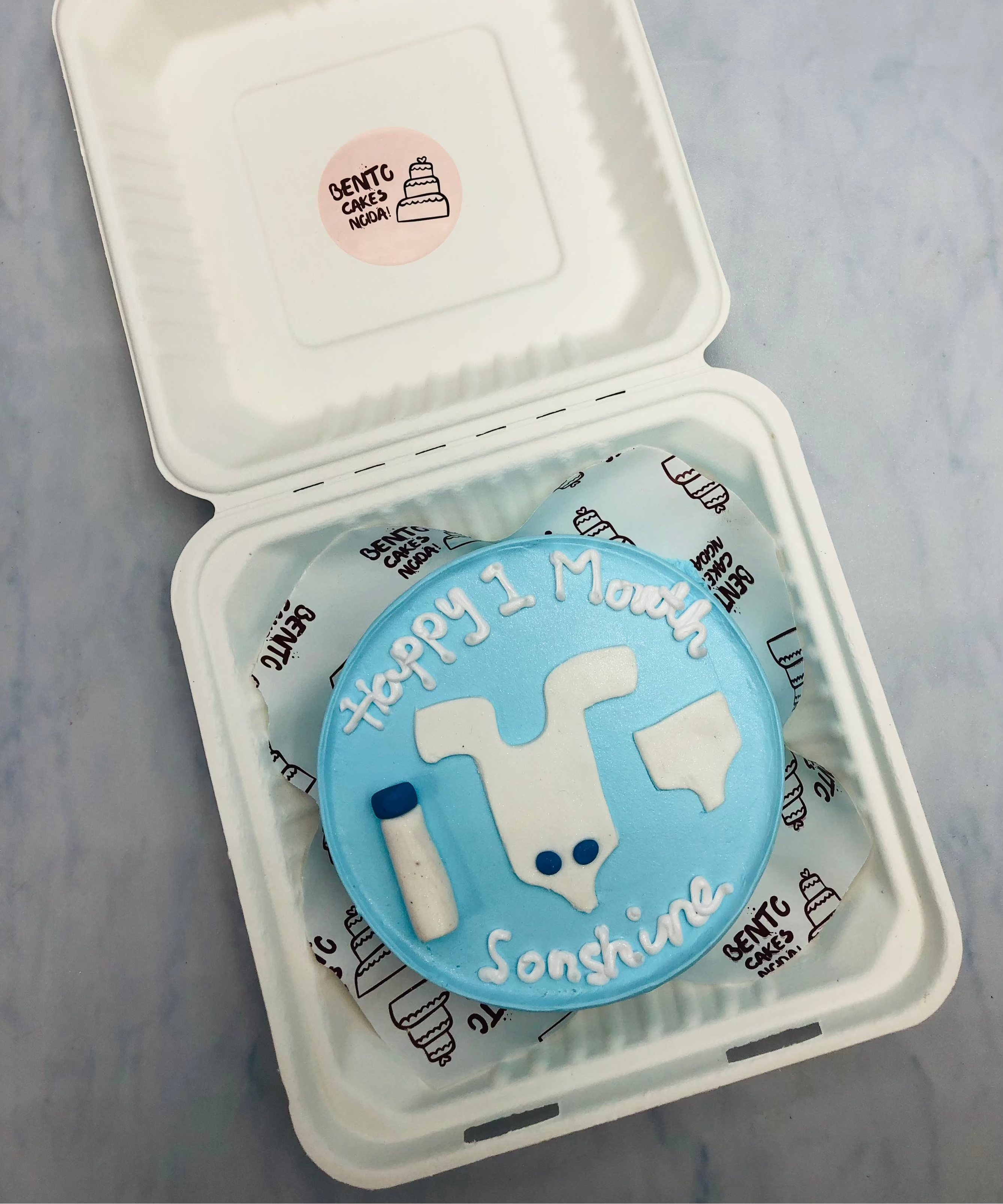 A sky blue bento cake with baby clotths and bottle design on it.
