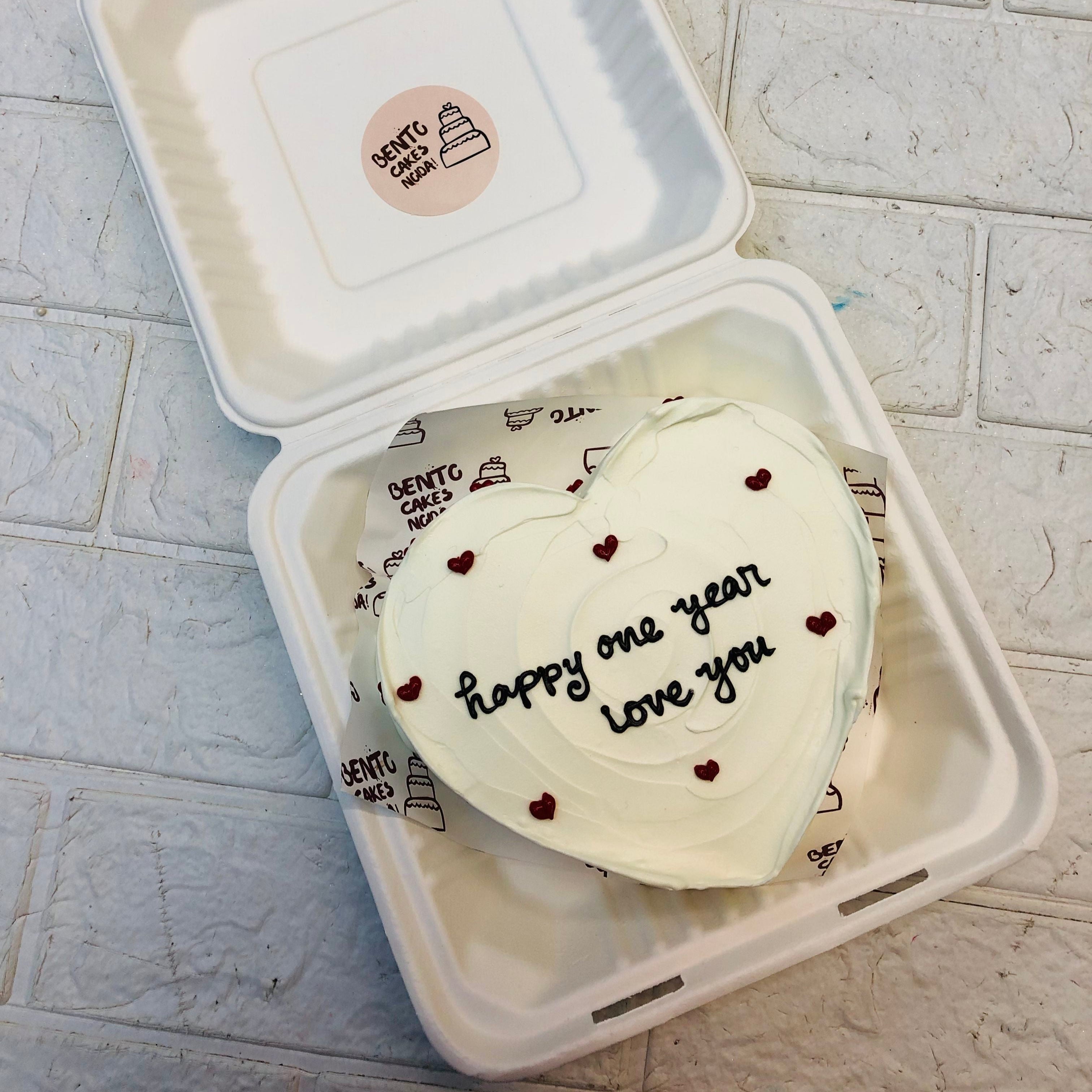 1st Anniversary white color heart shape bento cake with small red hearts and a loving text on it with blackm icing.