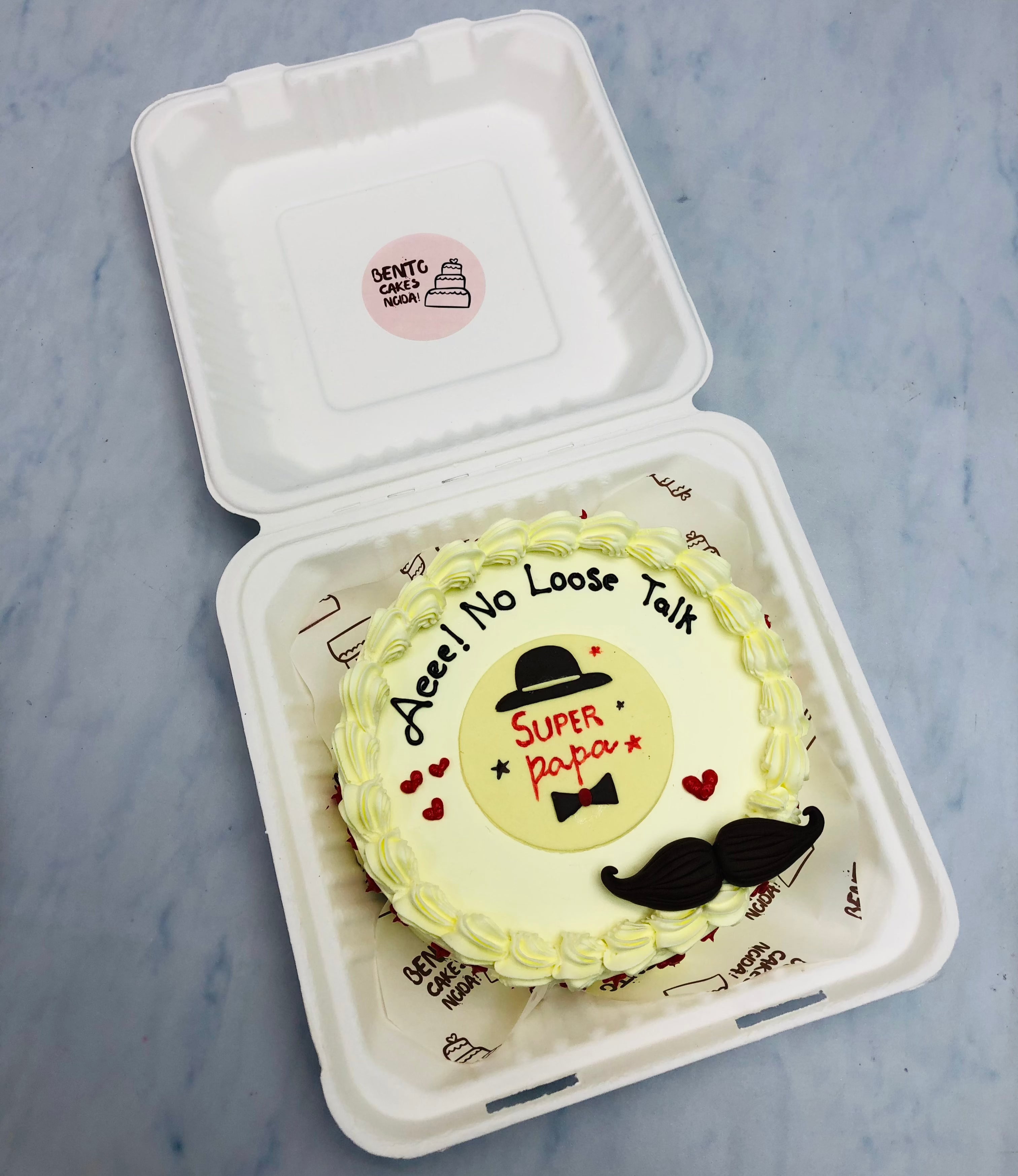 A bento cake with a "Super Papa" design is displayed in a white container. The cake features a yellow frosting border and is decorated with a black hat, mustache, and red hearts. The text "Aeee! No Loose Talk" is written on the cake, along with "Super papa" in the center.