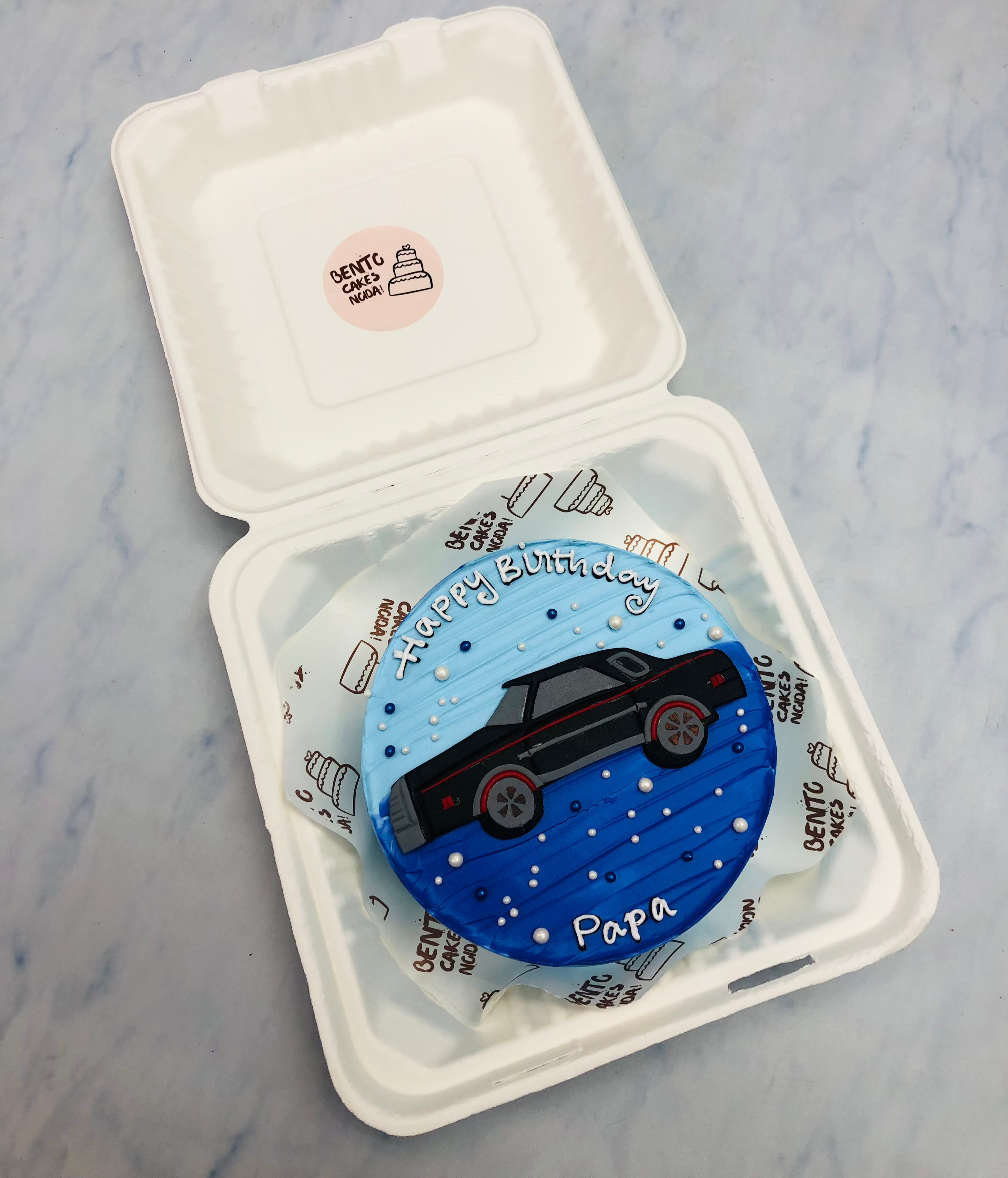 A round, personalized birthday cake featuring a stylish black car design on a blue gradient background, carefully placed inside a white takeaway box. The cake is inscribed with "Happy Birthday" at the top and "Papa" at the bottom in white icing. 