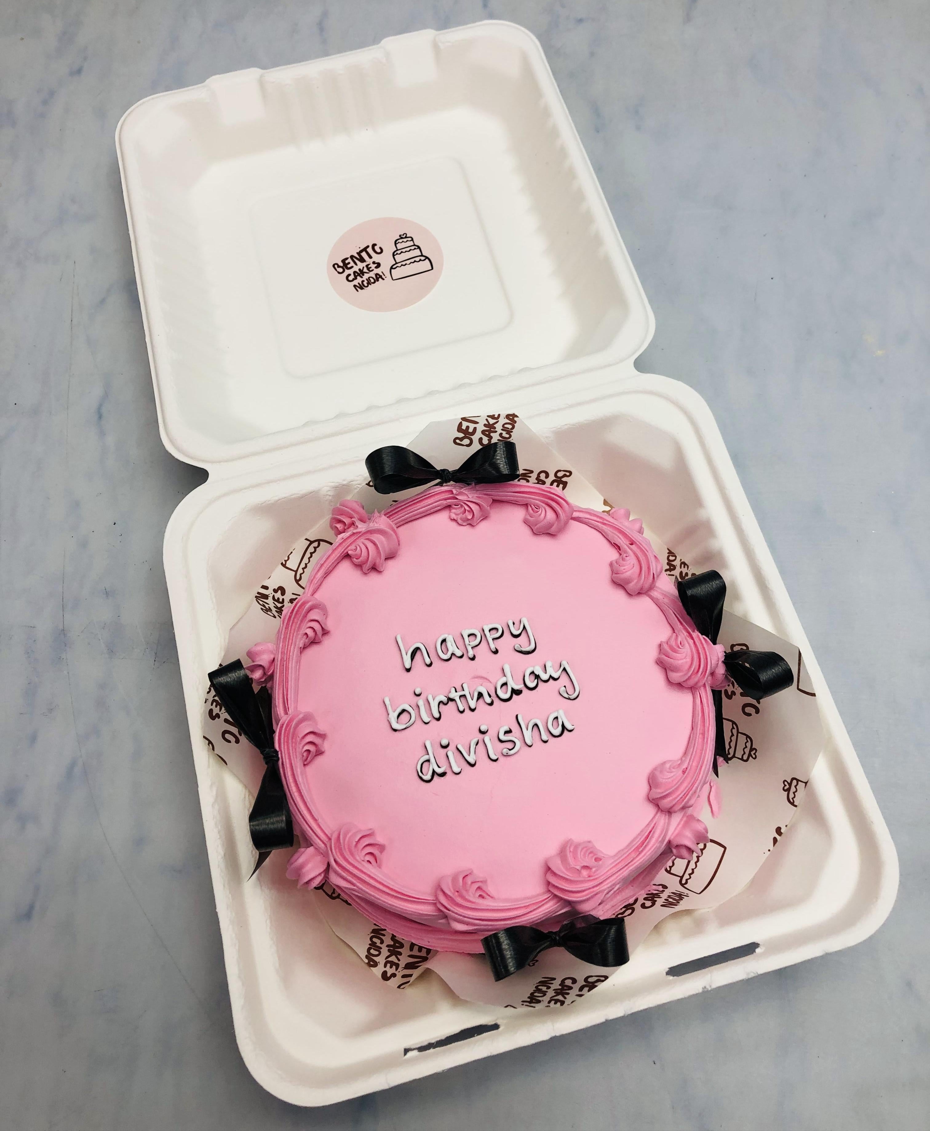 A pink birthday cake with the message "happy birthday divisha" written in white icing, placed inside a white bento box. The cake is decorated with pink frosting swirls along the edges and black ribbon bows around the sides.