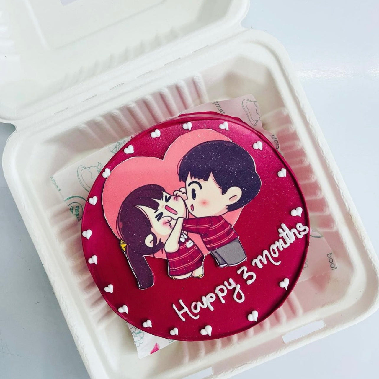 A round cake with a red icing base, featuring a cute cartoon illustration of a boy and girl in matching red outfits. The boy is playfully touching the girl's nose, and both are smiling. The cake is decorated with small white heart shapes around the edge and the text "Happy 3 months" written in white icing.
