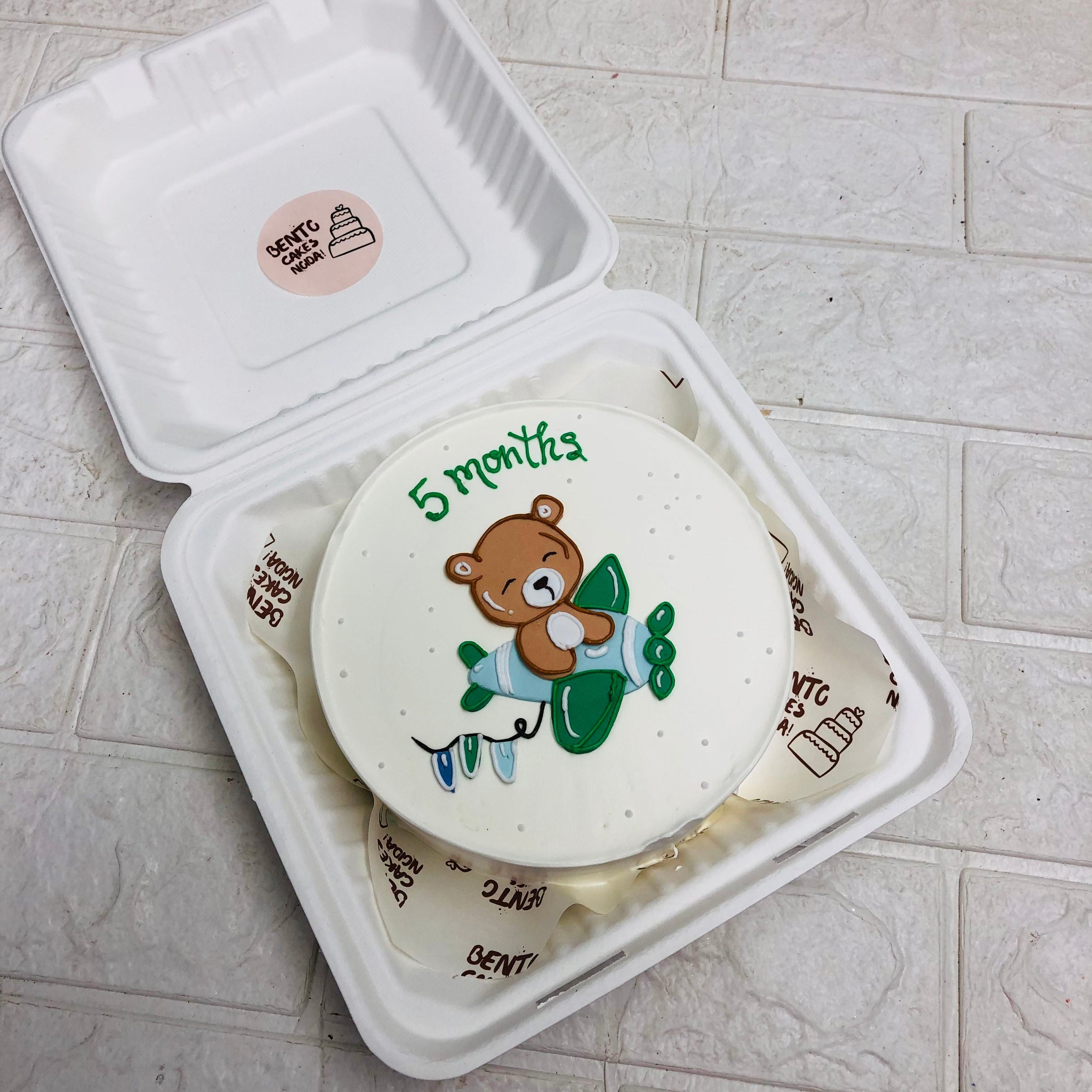 A white cake with a bear and airplane design, celebrating 5 months, placed in a white bento box.