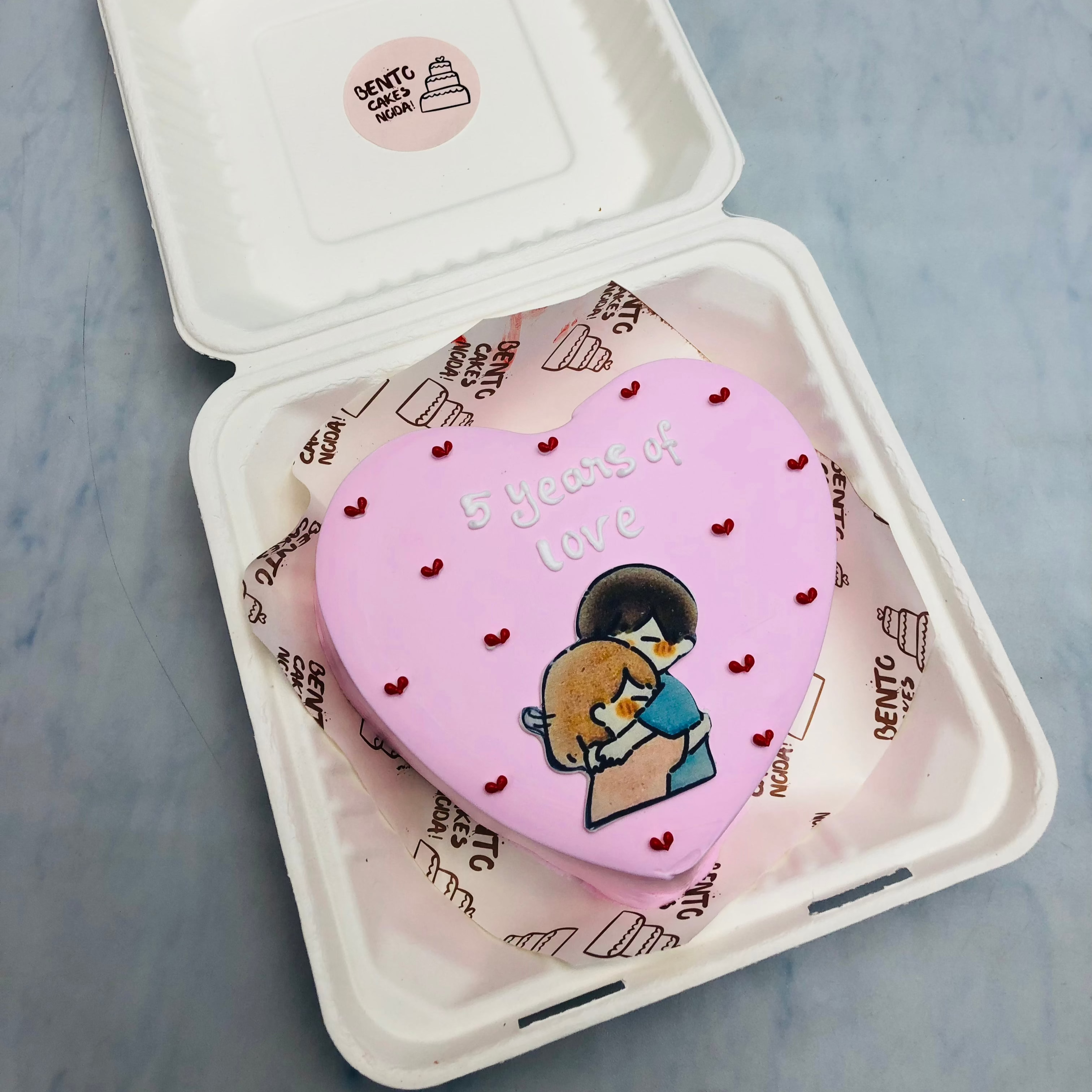 5 Years Love Theme Bento Cake withn light pink color and a hugging couple print on it with little red hearts all over the cake.