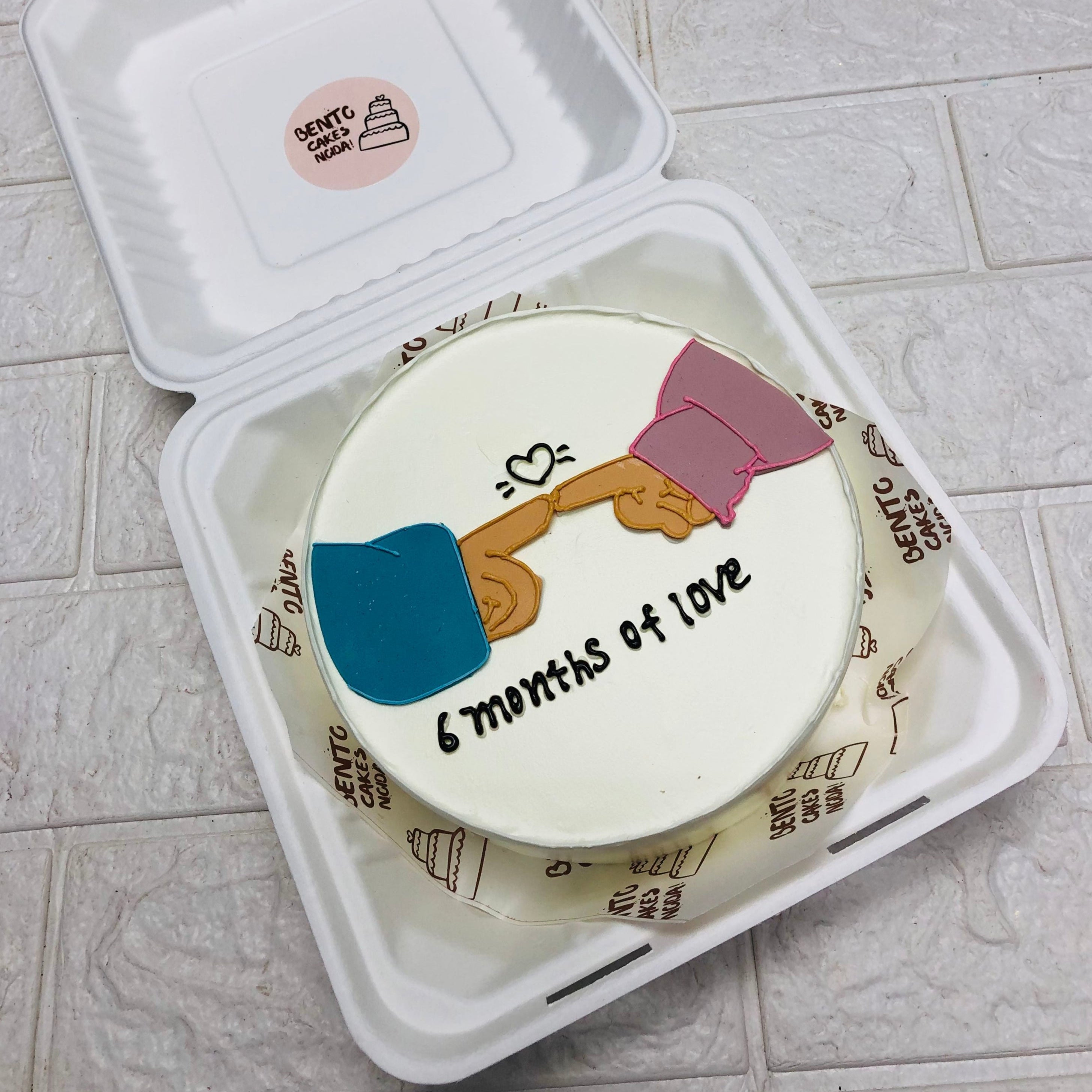 A round cake inside a white takeout box, featuring a design of two hands touching fingers (one in a blue sleeve and the other in a pink sleeve) with a heart above. The text on the cake reads "6 months of love".