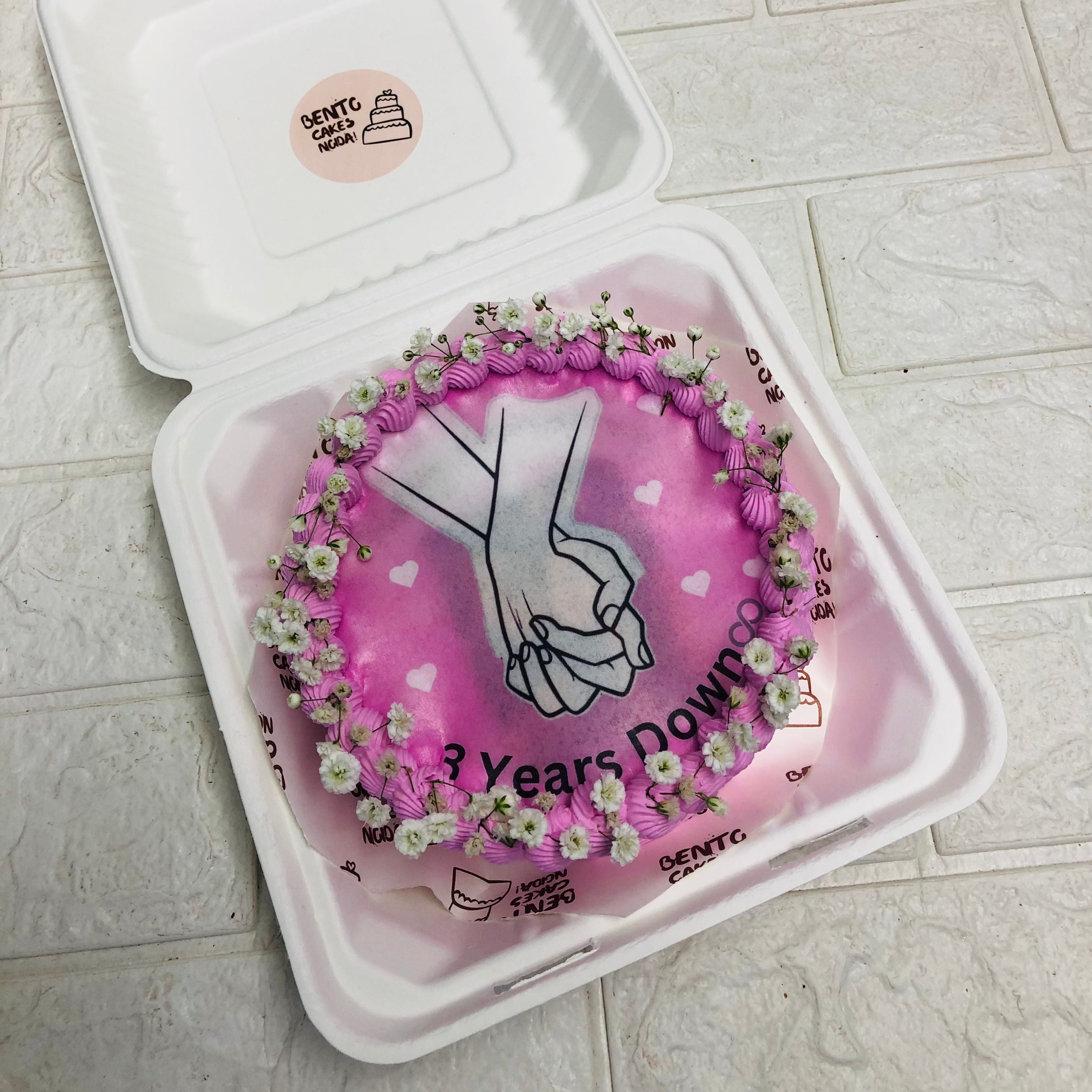  A decorative box featuring a cake inside, embellished with a pink heart with surrounded by flowers, representing love and special occasions.