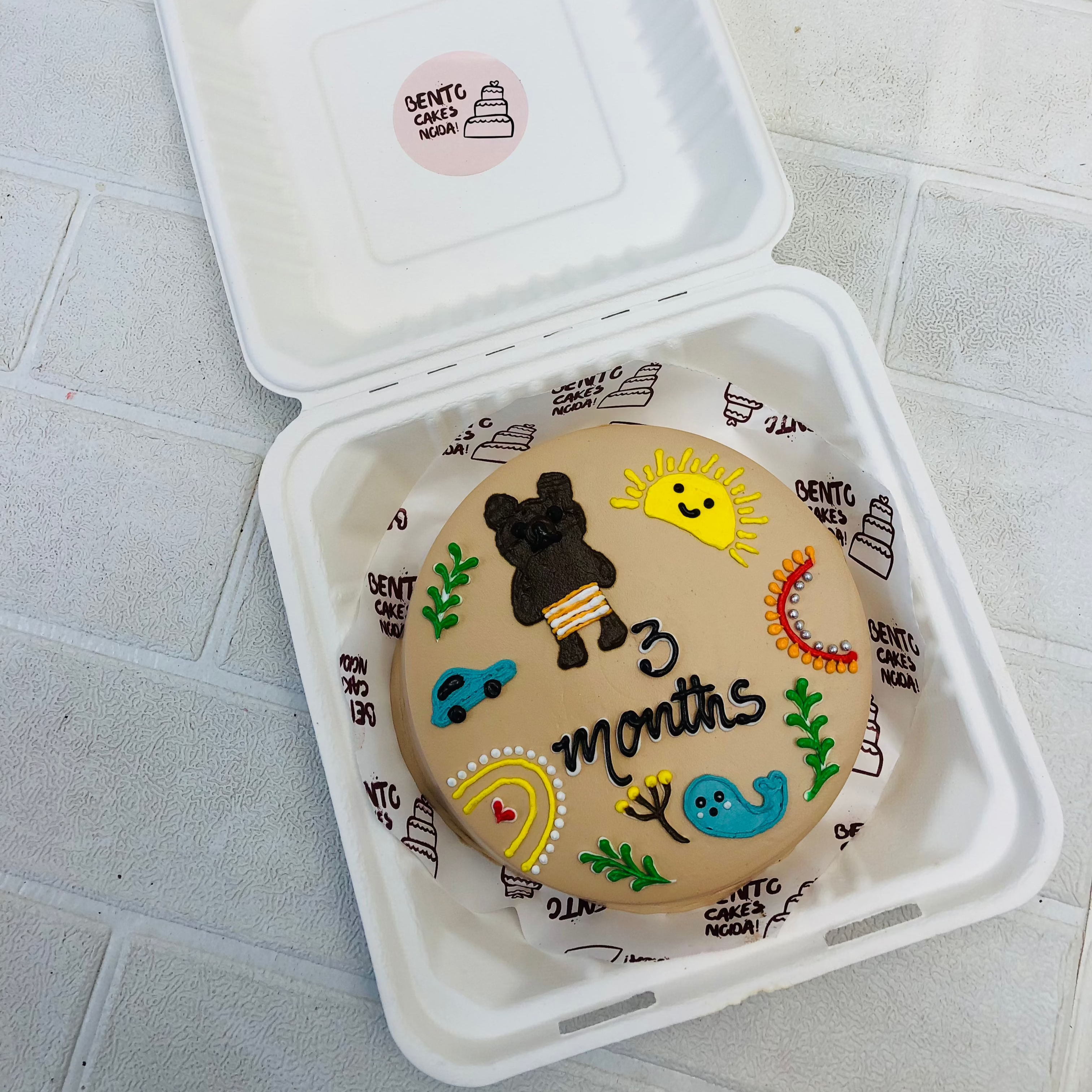 A light brown bento cake with cute brown teddy , car, fish, son etc drawings on cake.