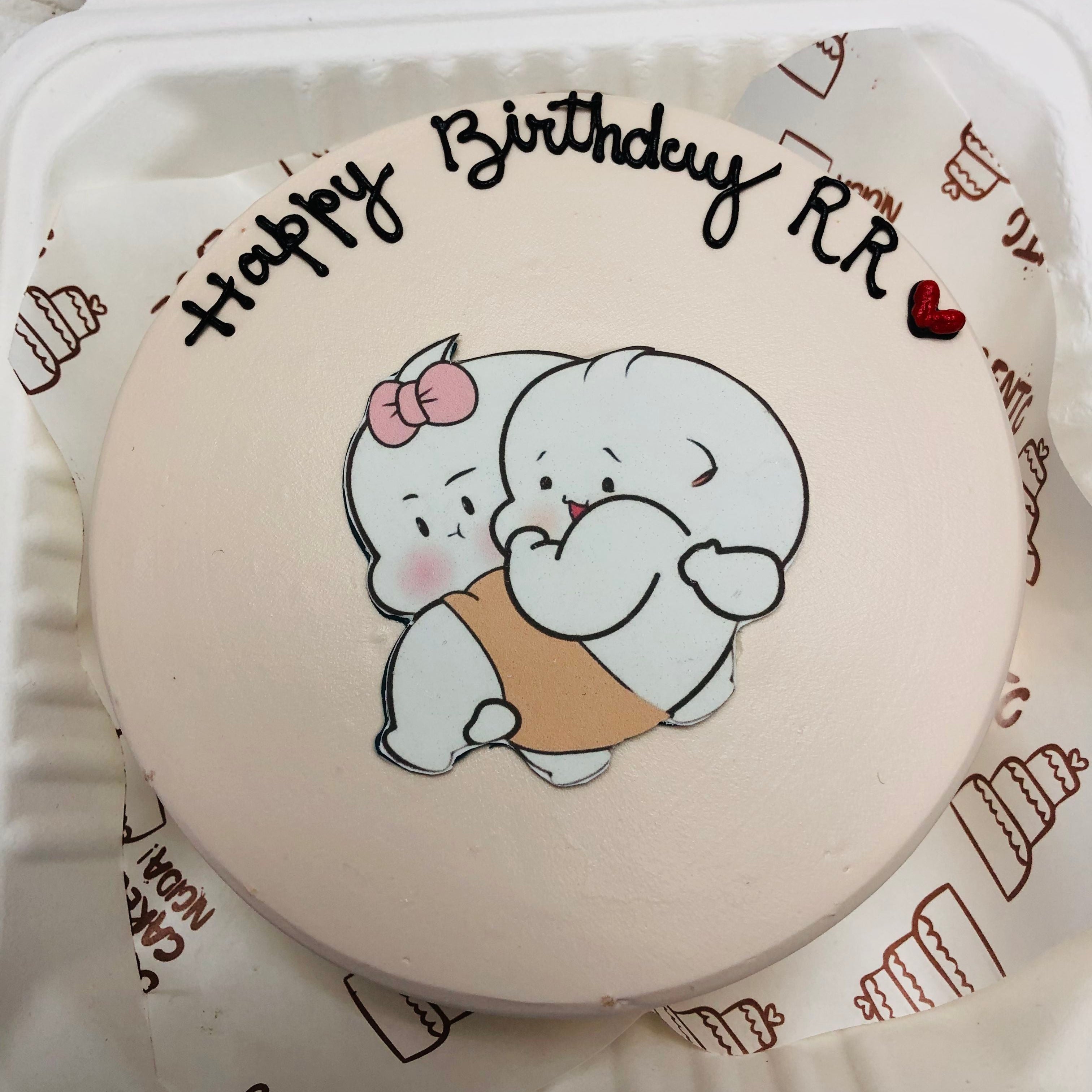 A round birthday cake with a light pink frosting, featuring a cute cartoon illustration of two white characters hugging, one with a pink bow. The cake is inscribed with 'Happy Birthday RR'.