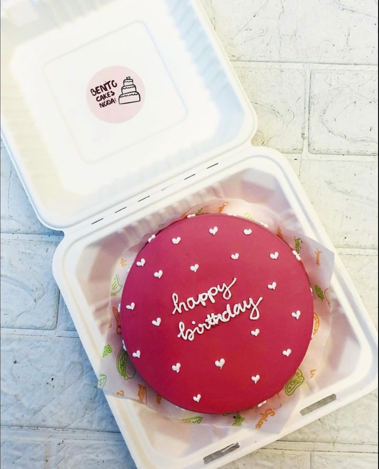 A dark red bento cake is designed with little white hearts design and a text "happy birthday" written with white icing.