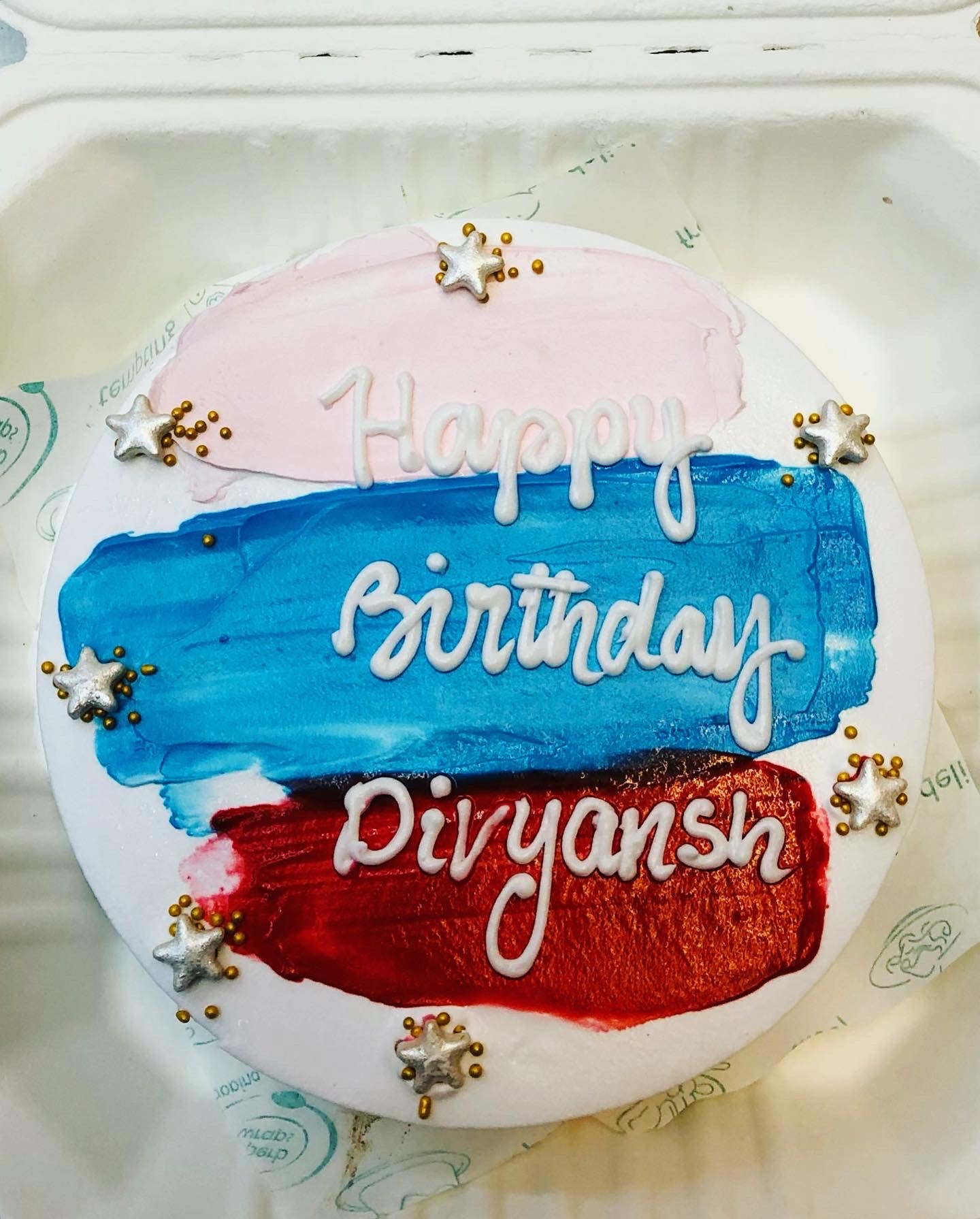 A round birthday cake with pink, blue, and red frosting, decorated with silver star-shaped sprinkles and a personalized message that reads 'Happy Birthday Divyansh'.