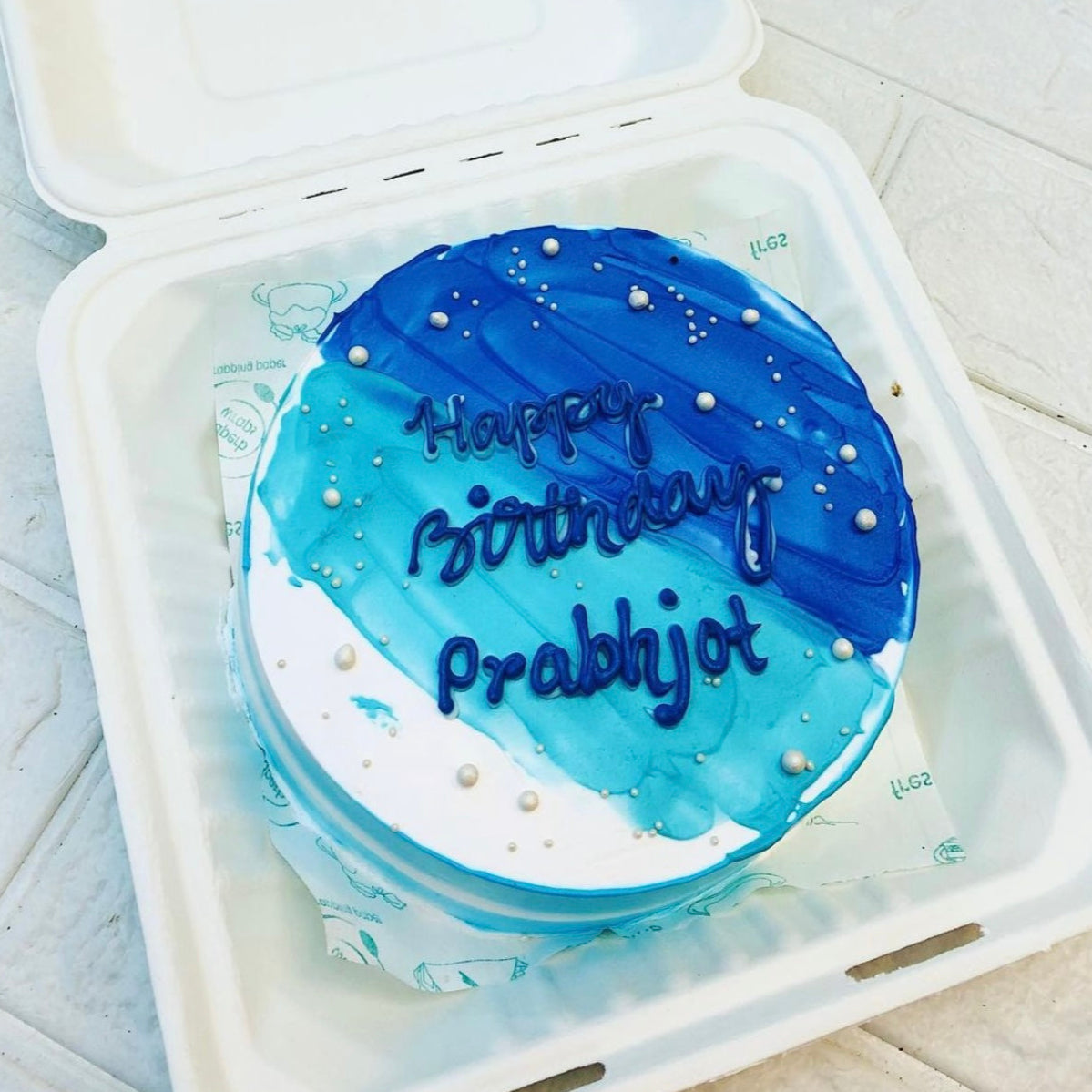 A round birthday cake with blue and white frosting, decorated with small white pearls, and the message "Happy Birthday Prabhjot" written in blue icing.
