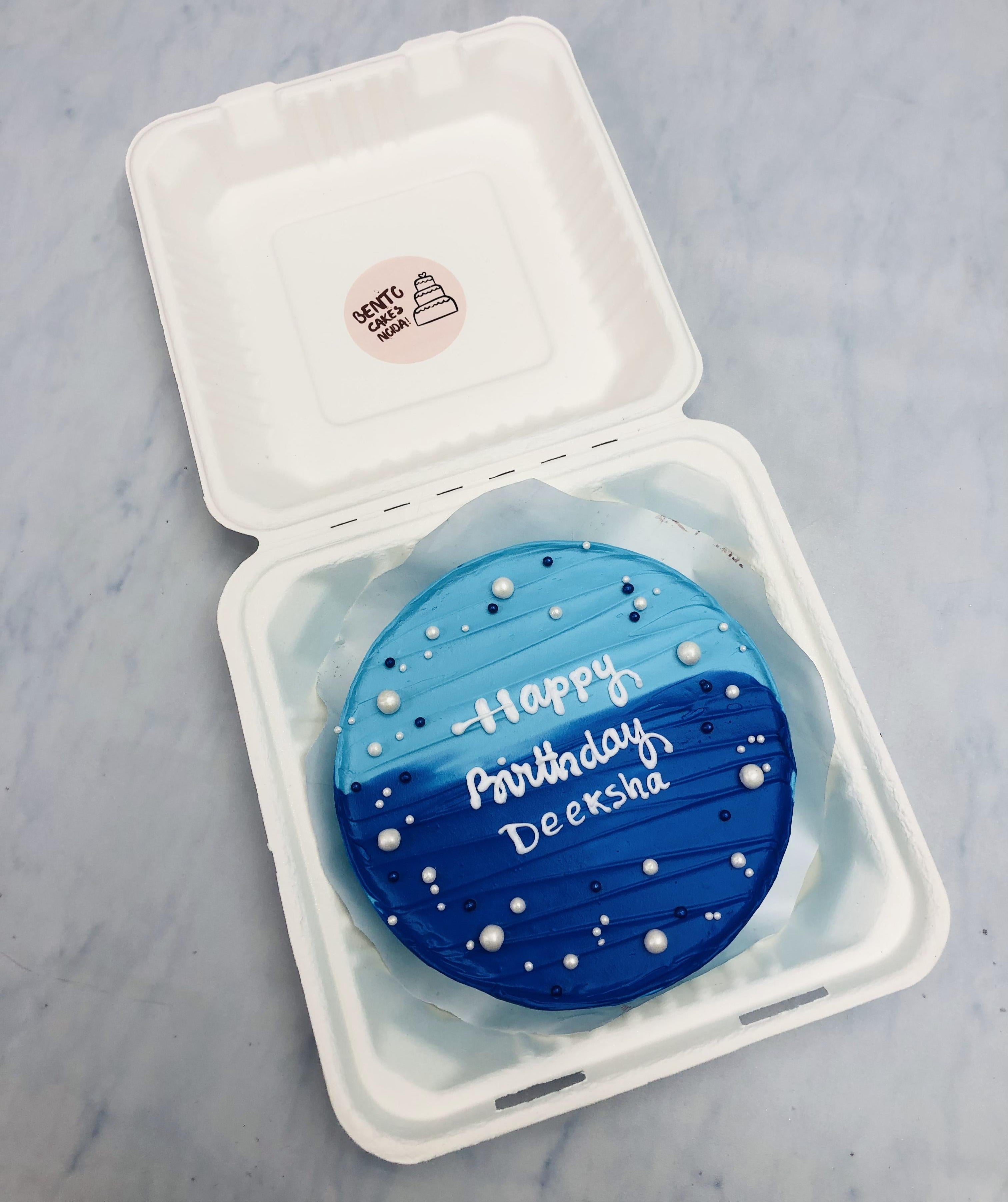 An aesthetic blue frosting bento cake with 'happy birthday Deeksha' written in white icing, placed inside a white bento box. The cake is decorated with small white and blue sprinkles.