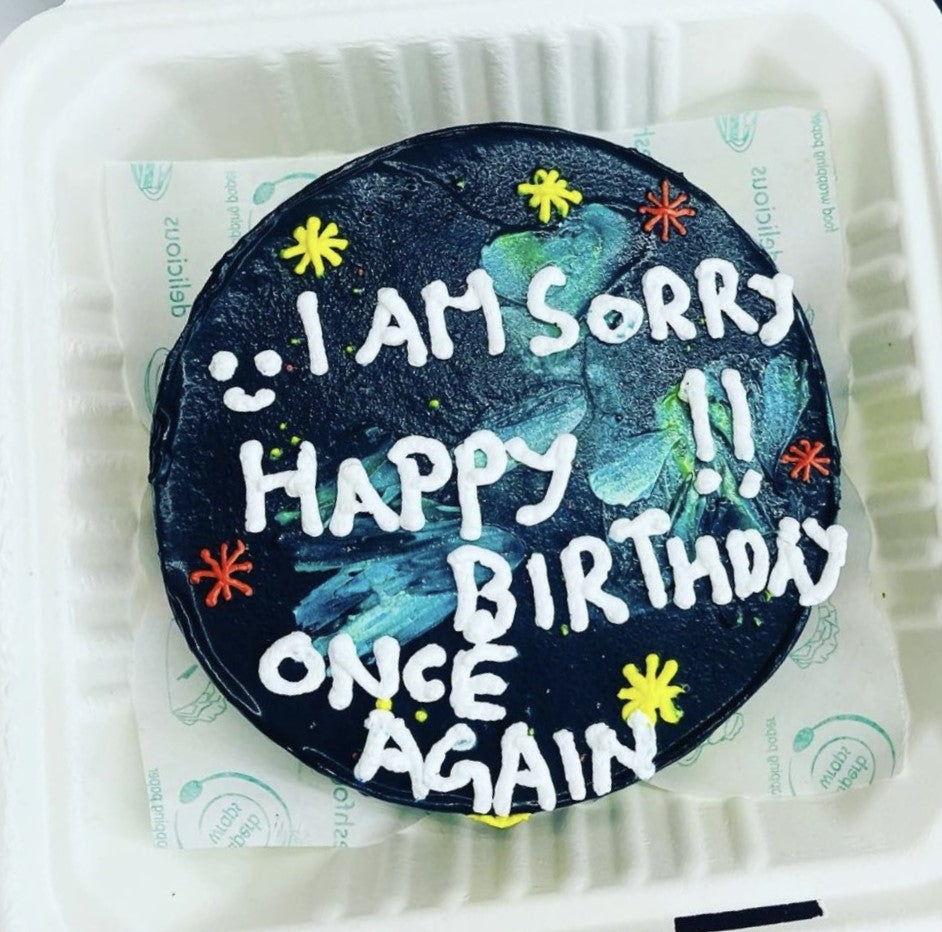 A round cake with black frosting, decorated with colorful star-shaped sprinkles and white icing text that reads, "I AM SORRY HAPPY BIRTHDAY ONCE AGAIN," placed in a white container.