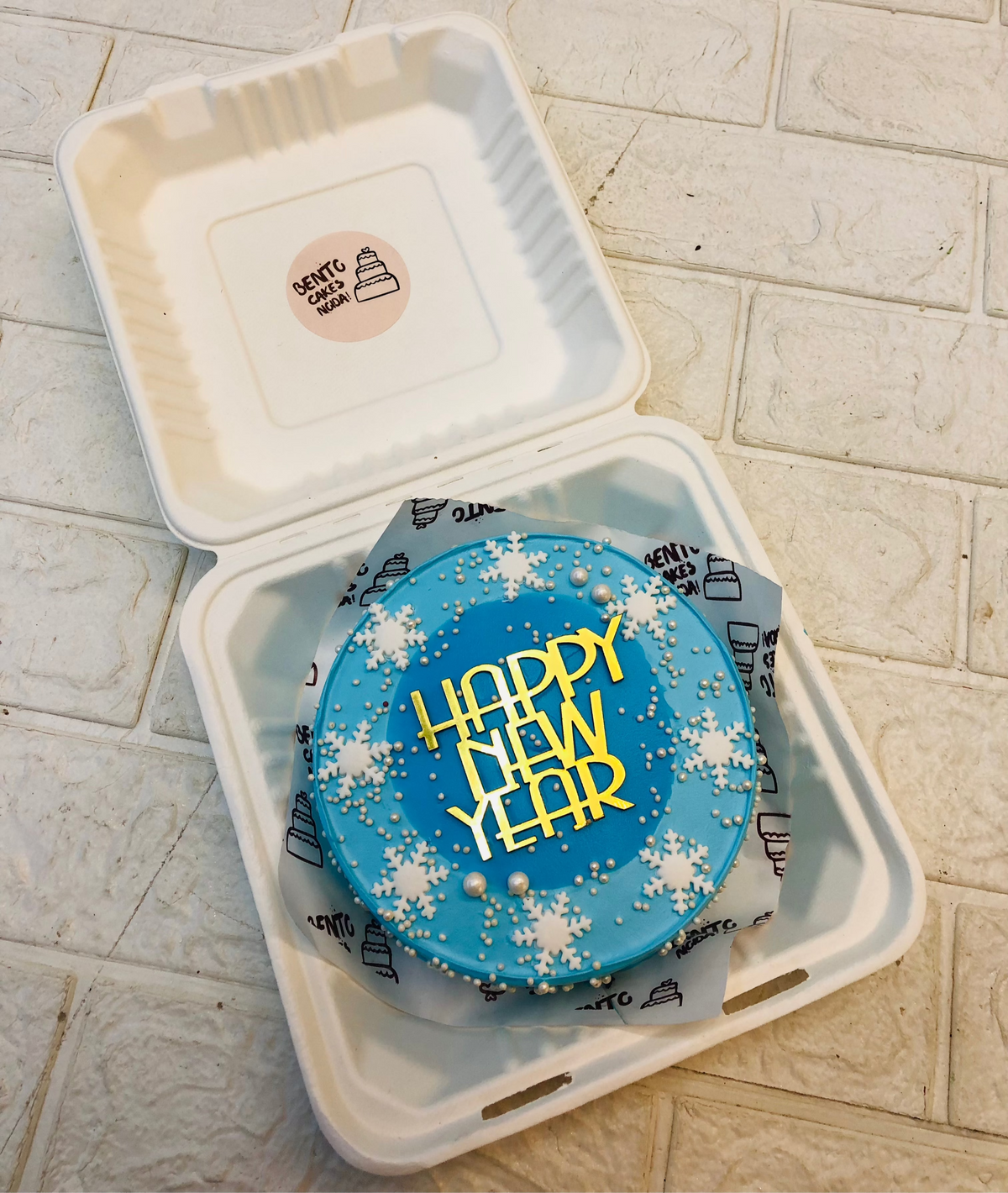 A skyblue bento cake with dark blue circular design on its center and a golden topper of Happty New Year placed on it.