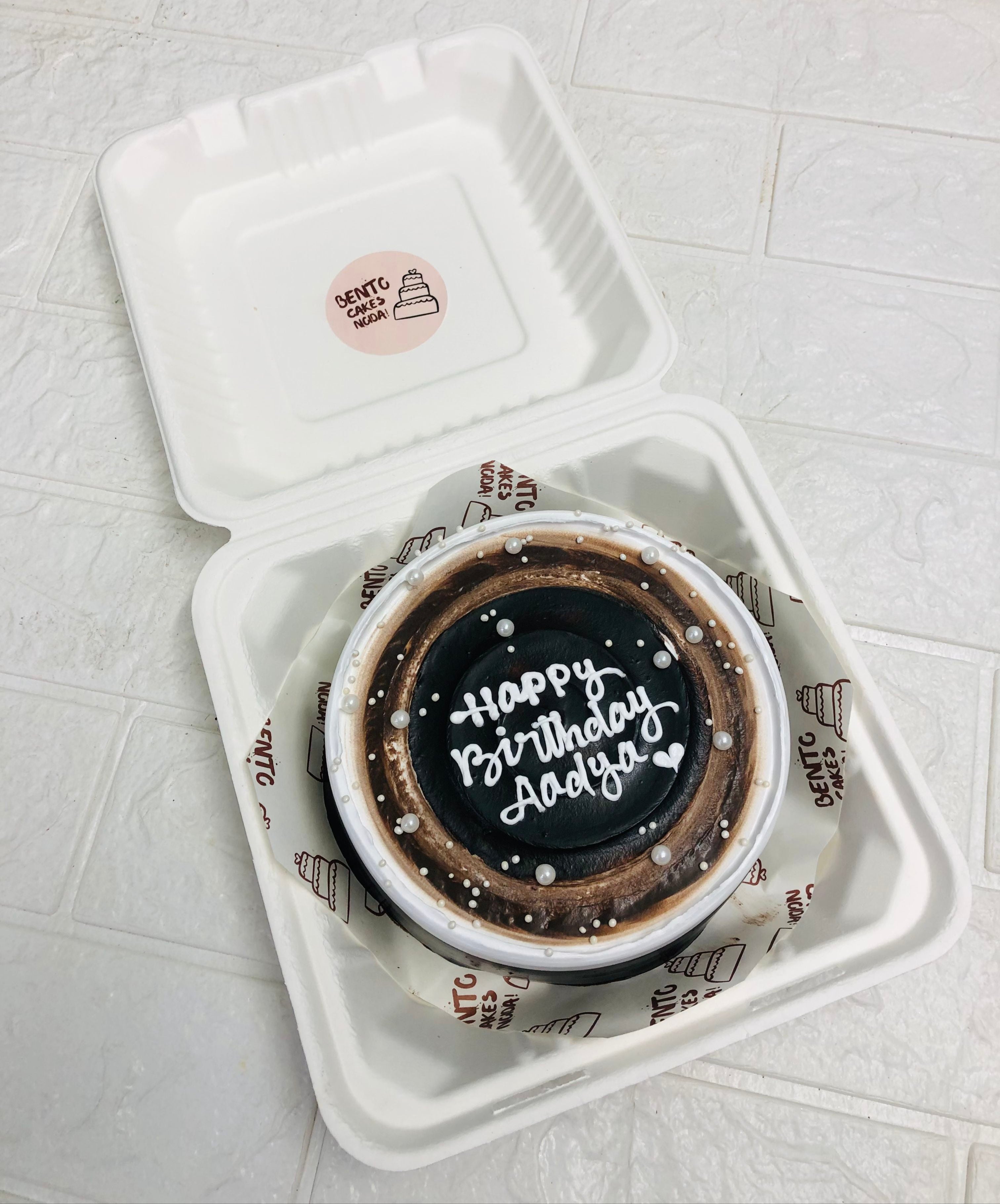 A beautifully decorated birthday cake with the message "Happy Birthday Dadya" written in white icing. The cake is placed inside a bento box. It features a gradient design with shades of brown and white, adorned with small white edible pearls.