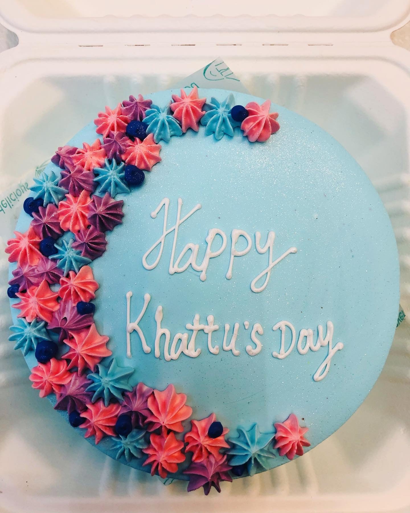 A round blue cake with the words 'Happy Khattu's Day' written in white icing. The cake is decorated with an array of colorful star-shaped frosting in shades of pink, purple, and blue, arranged in a crescent shape on the left side.