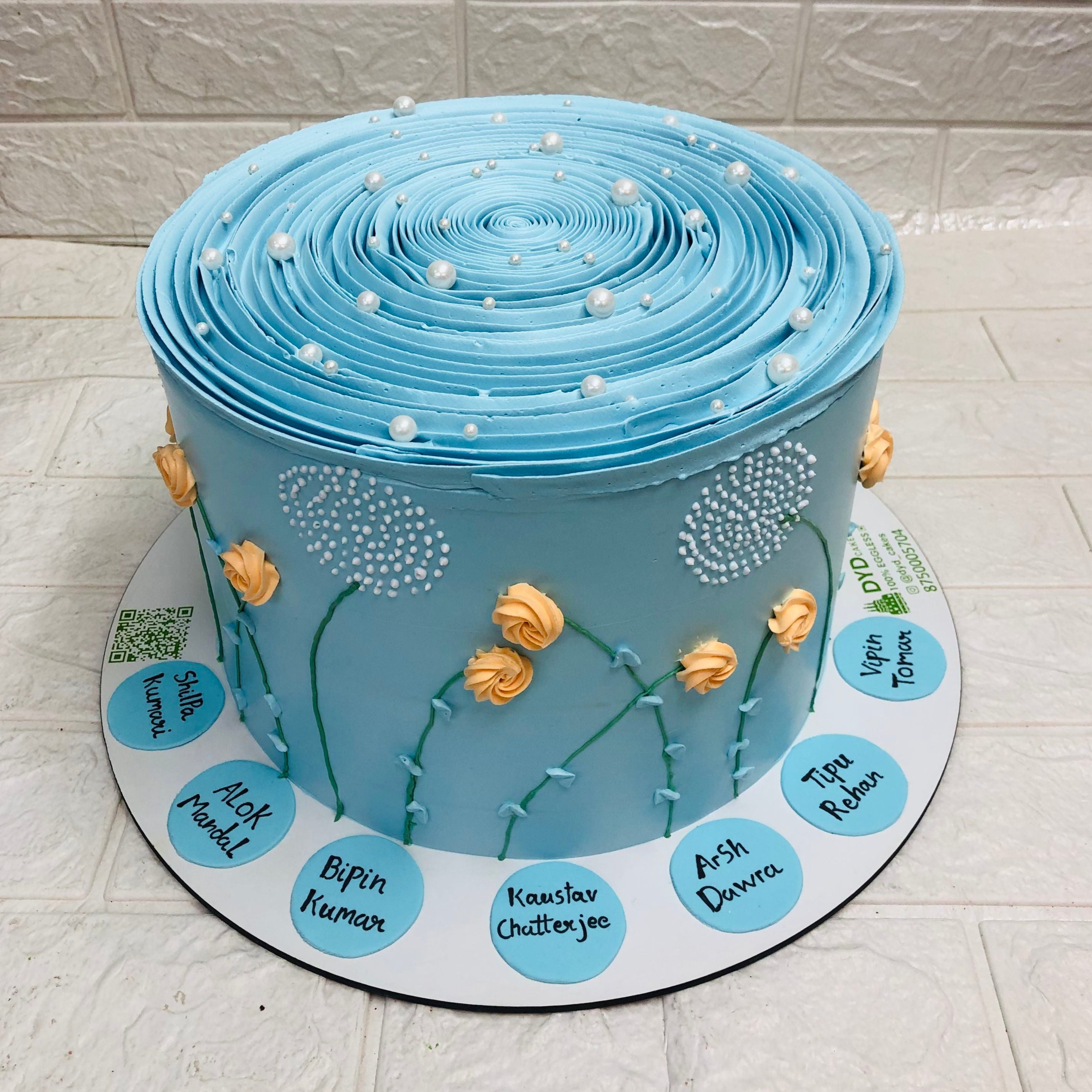 A stunning blue-frosted cake featuring intricate floral designs and pearl-like embellishments. The cake showcases yellow rose-like decorations and delicate white patterns resembling dandelions. Placed on a white base, it includes blue circles with handwritten names, adding a personal touch. Perfect for special celebrations and memorable occasions.