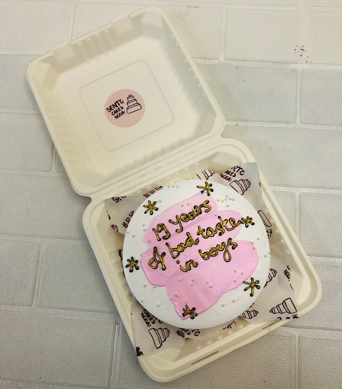 An aesthetic white bento cake with baby pink shades on top and decorated with golden stars and a golden text.