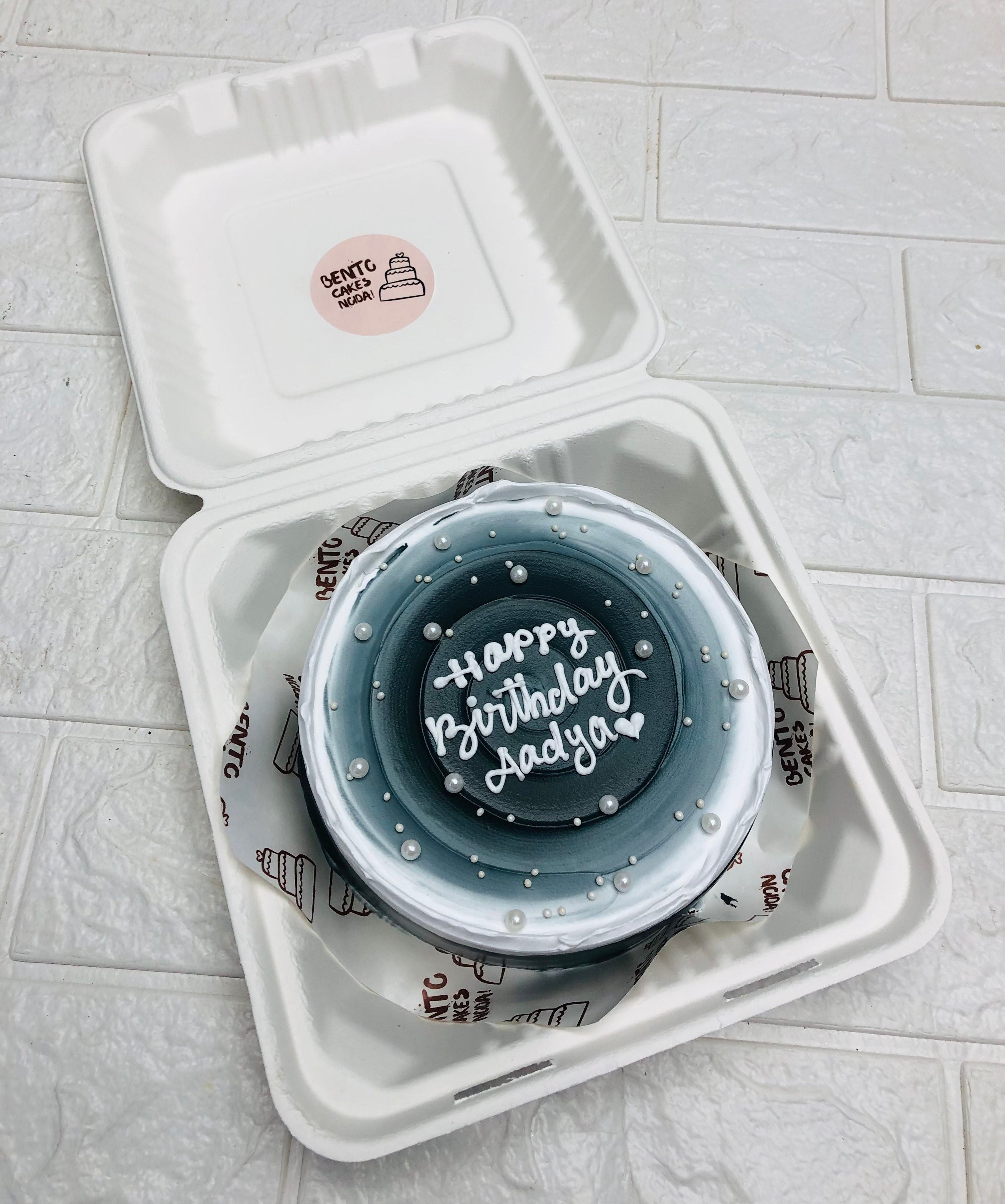 A beautifully decorated birthday cake with the message "Happy Birthday Aadya" written in white icing. The cake is placed inside an eco-friendly, white, biodegradable takeout box. It features a gradient design with shades of blue and white, adorned with small white edible pearls.