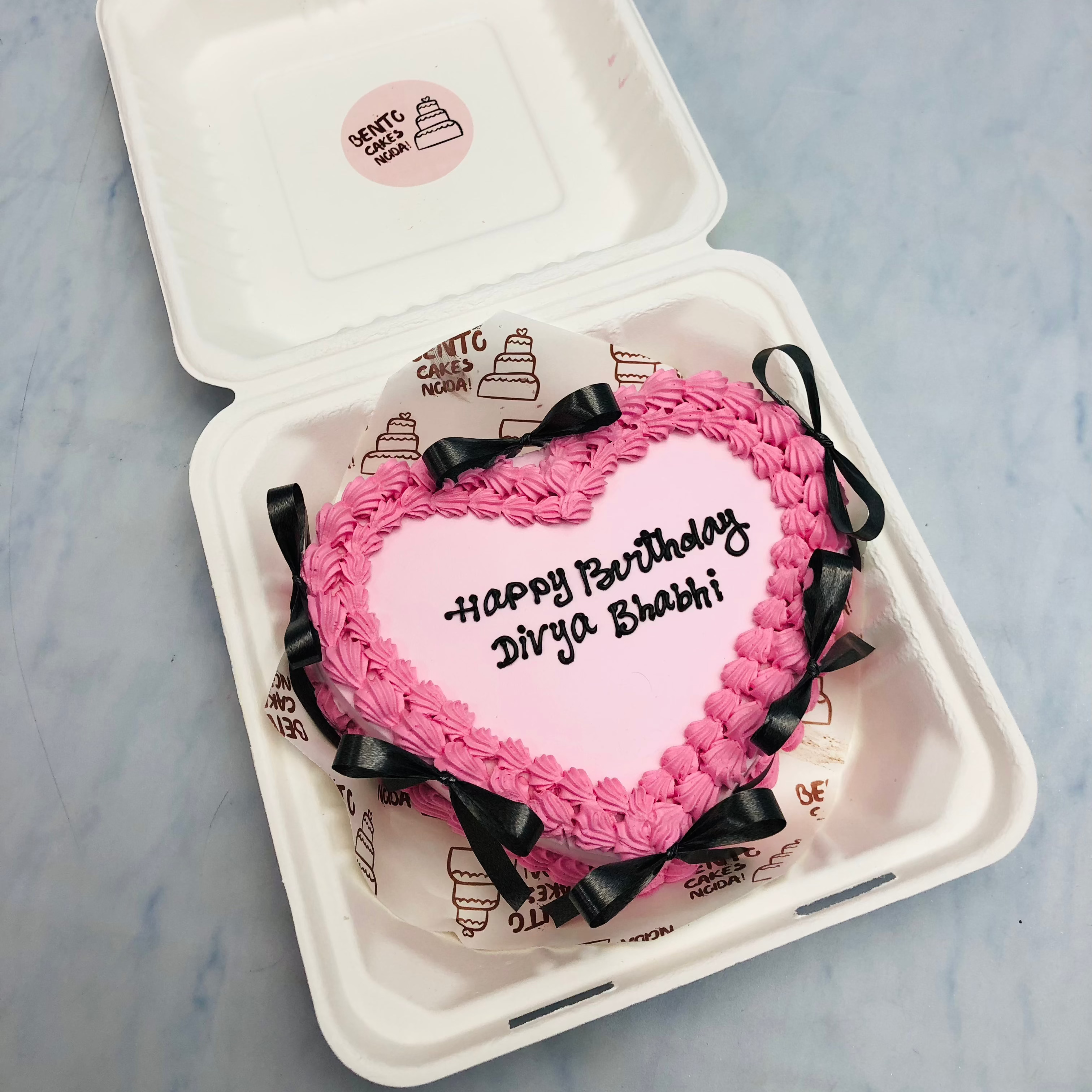 A light pink heart shape cake ith pink truffle boundary and decorated with black bows all over the cake.