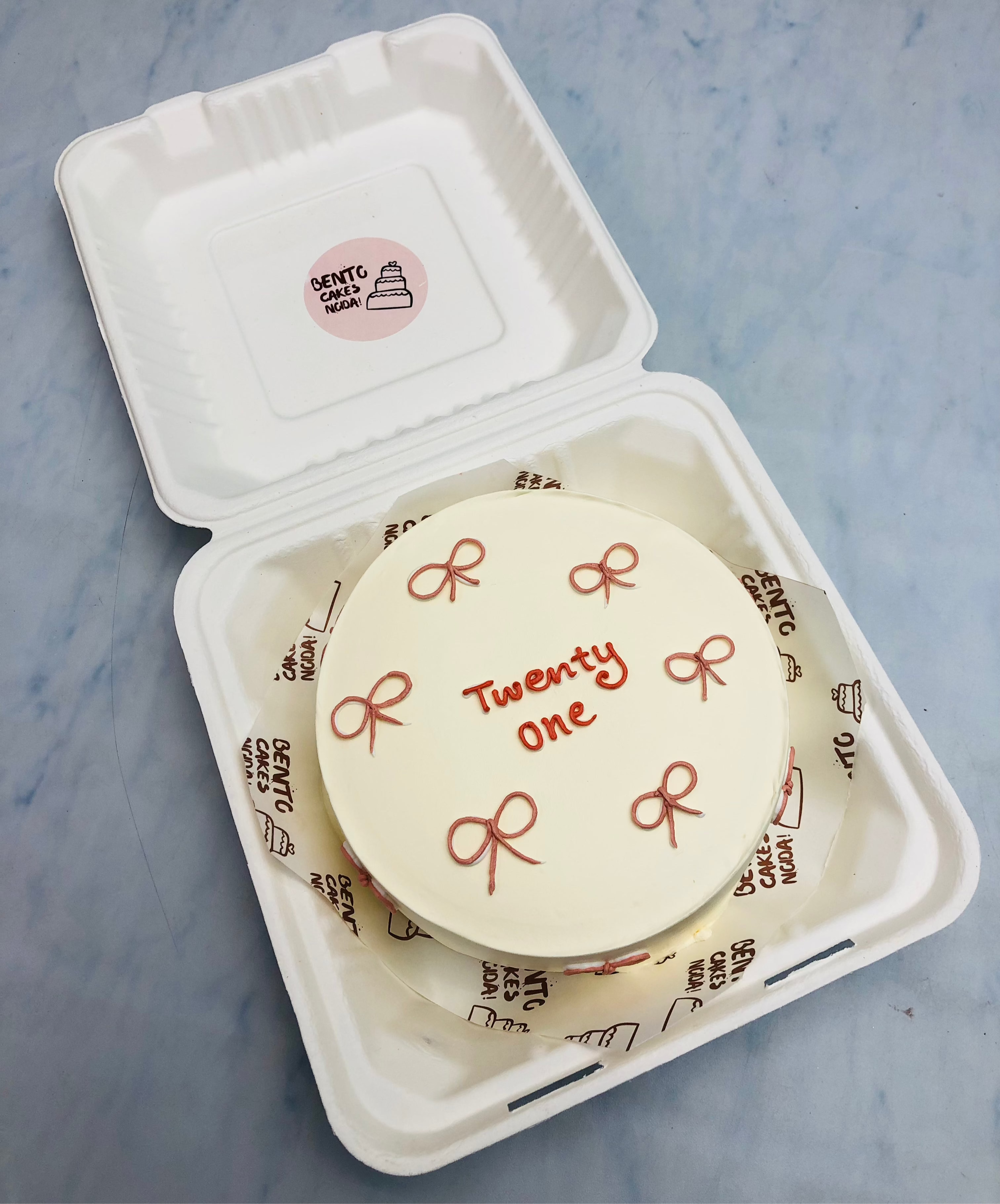 A round, minimalist white cake featuring “Twenty One” written in red icing at the center, accompanied by delicate red bow decorations.