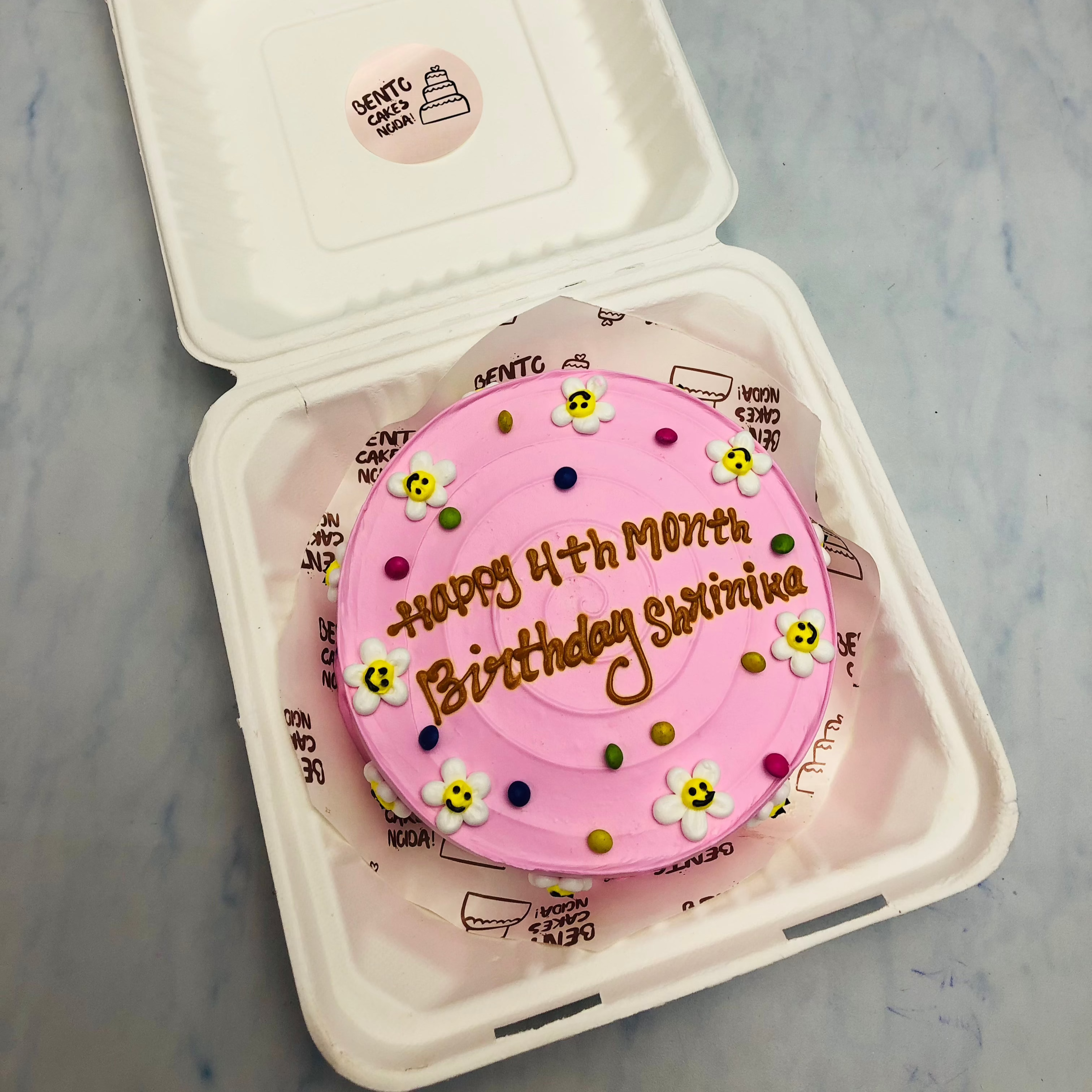 A light pink bento cake with cute floral designs and a text written with brown icing.