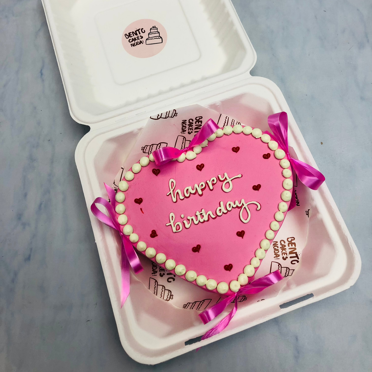 A pink color heart shape cake decorated with pink bow and boundries with white icing on it and having red heart design all over the cake.