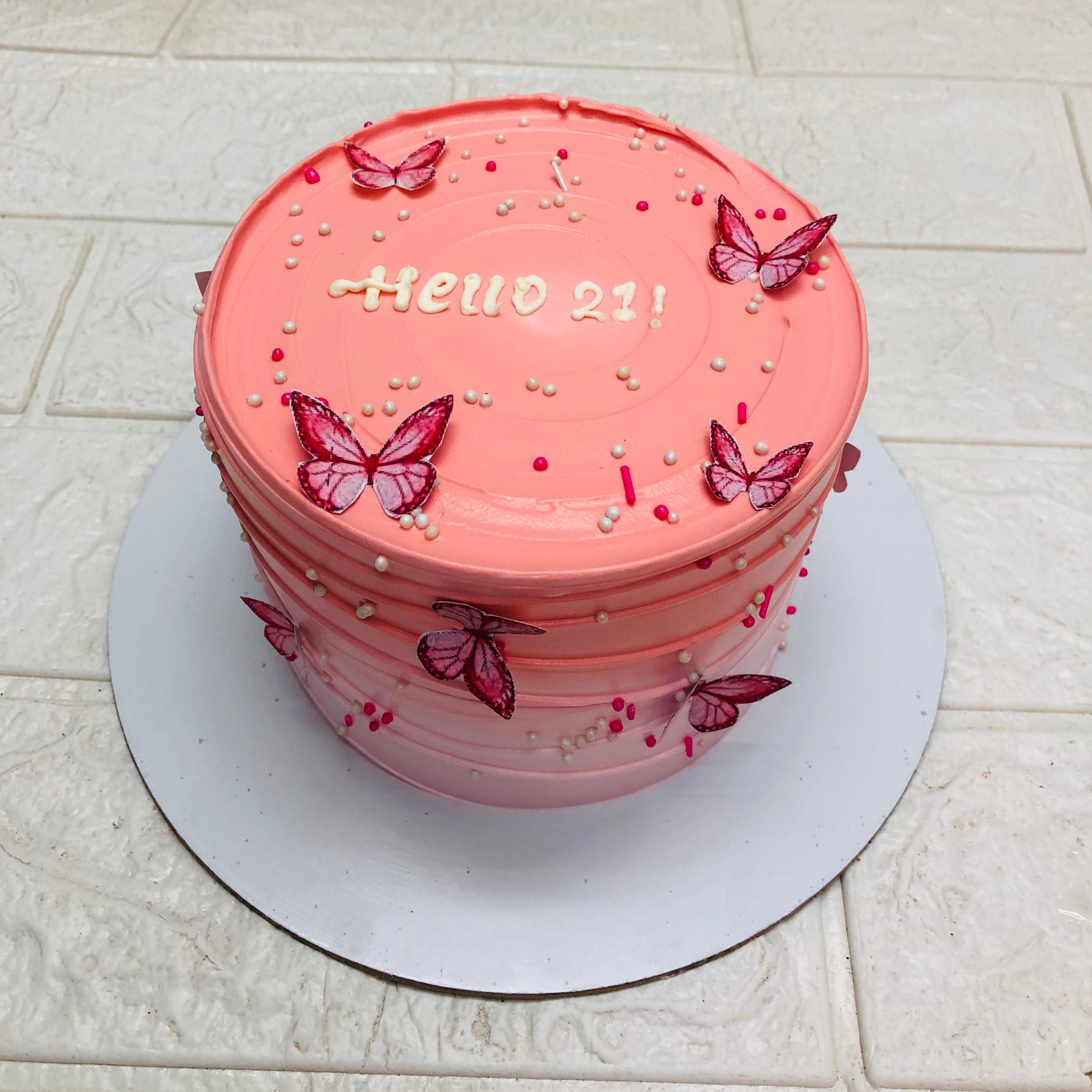 A beautifully decorated pink birthday cake with the text "Hello 21!" on top, adorned with pink butterfly decorations and colorful sprinkles.