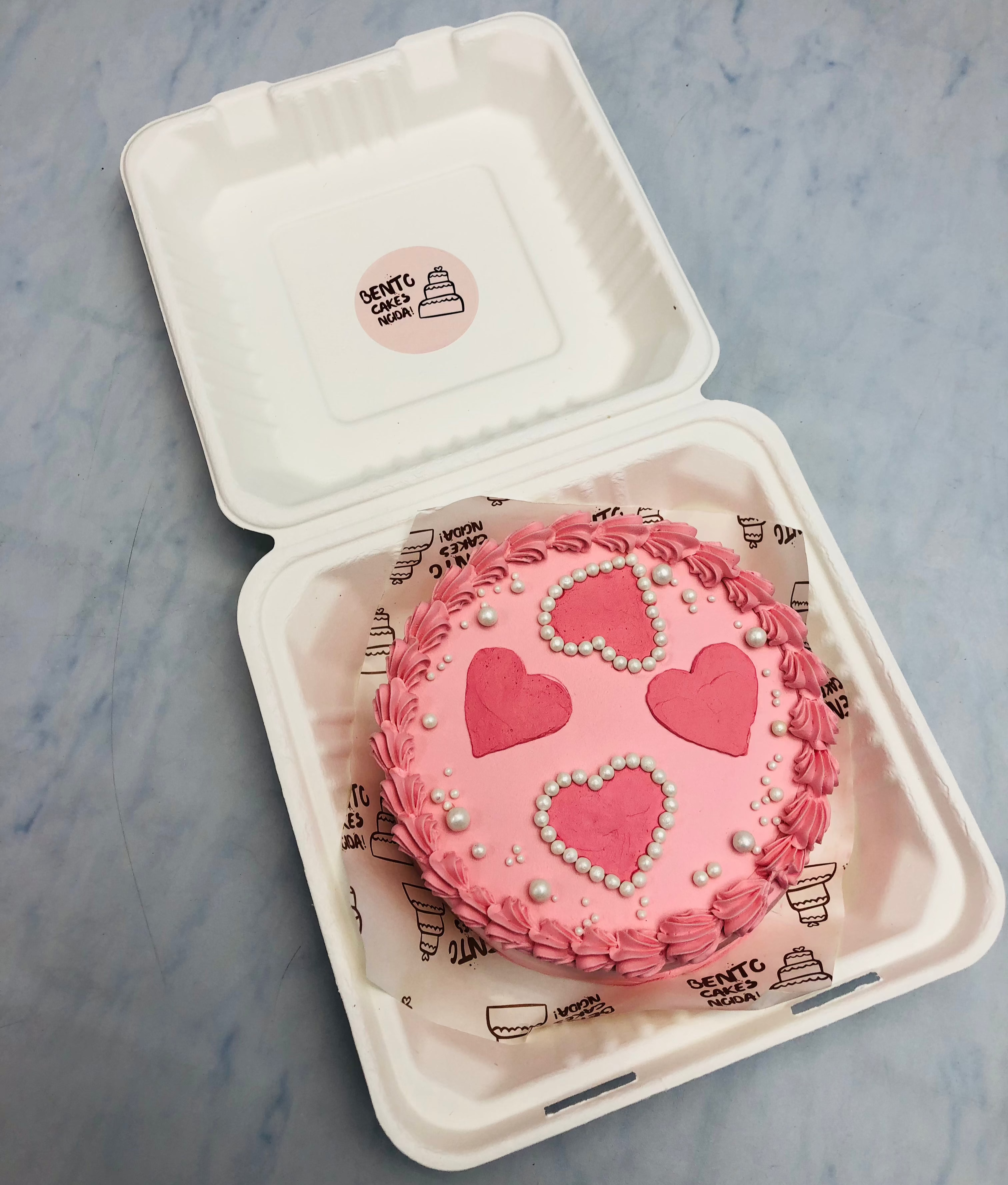 A charming pink bento cake adorned with three heart shapes and delicate pearl-like beads, nestled inside a sleek white eco-friendly container. The cake features a decorative pink frosting border.