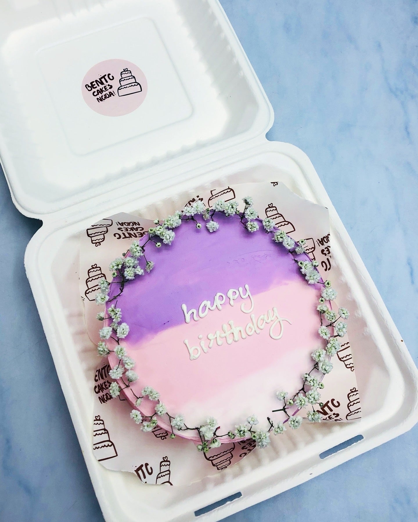 A beautifully decorated bento cake in a white container, featuring a soft gradient of purple and pink icing with delicate white flowers arranged in a ring around the edge. The cake has 'happy birthday' elegantly written in white icing, making it perfect for a special celebration.