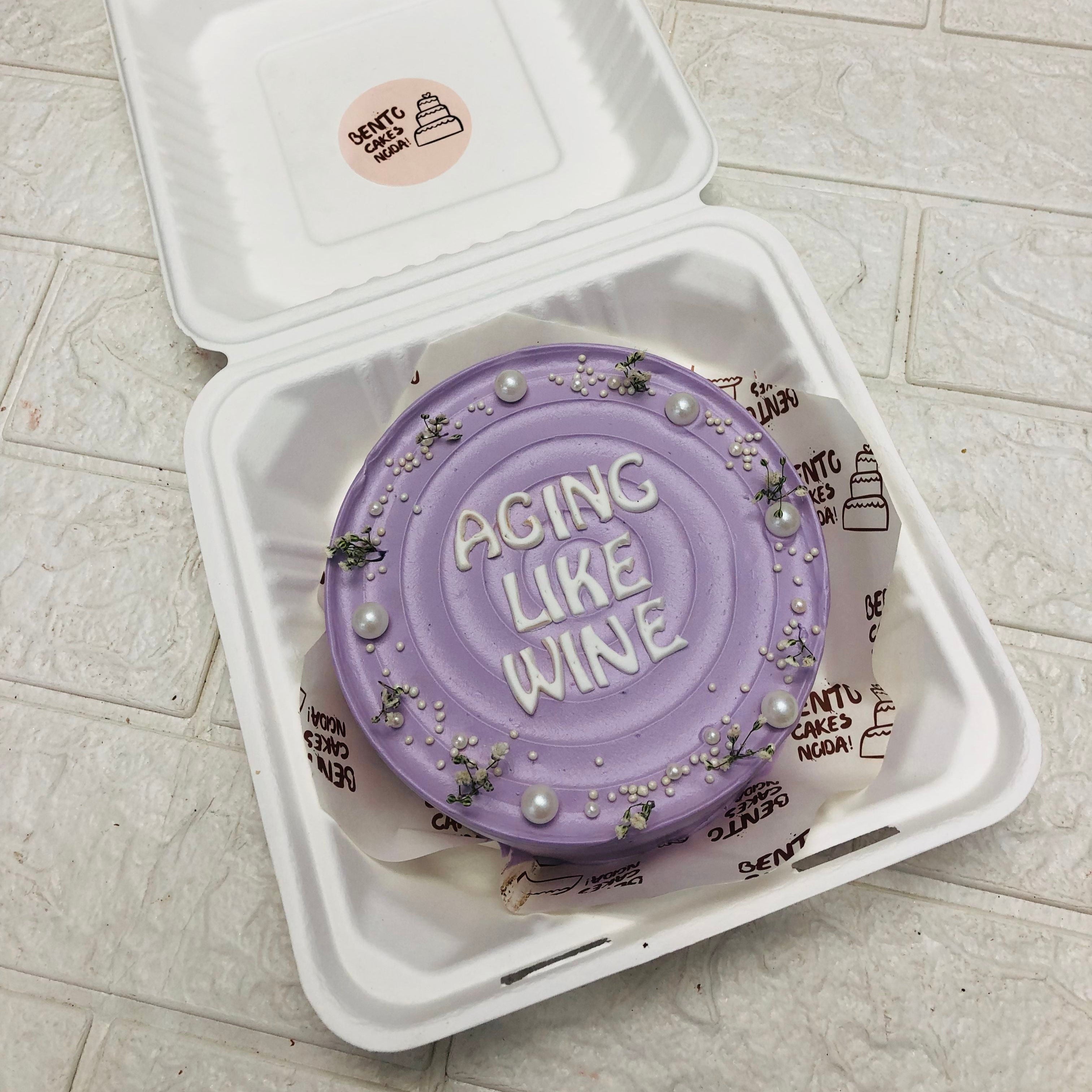 A small, round cake with light purple frosting, decorated with white pearls and small green leaves, placed inside a bento box. The cake has the words "AGING LIKE WINE" written in white icing on top.