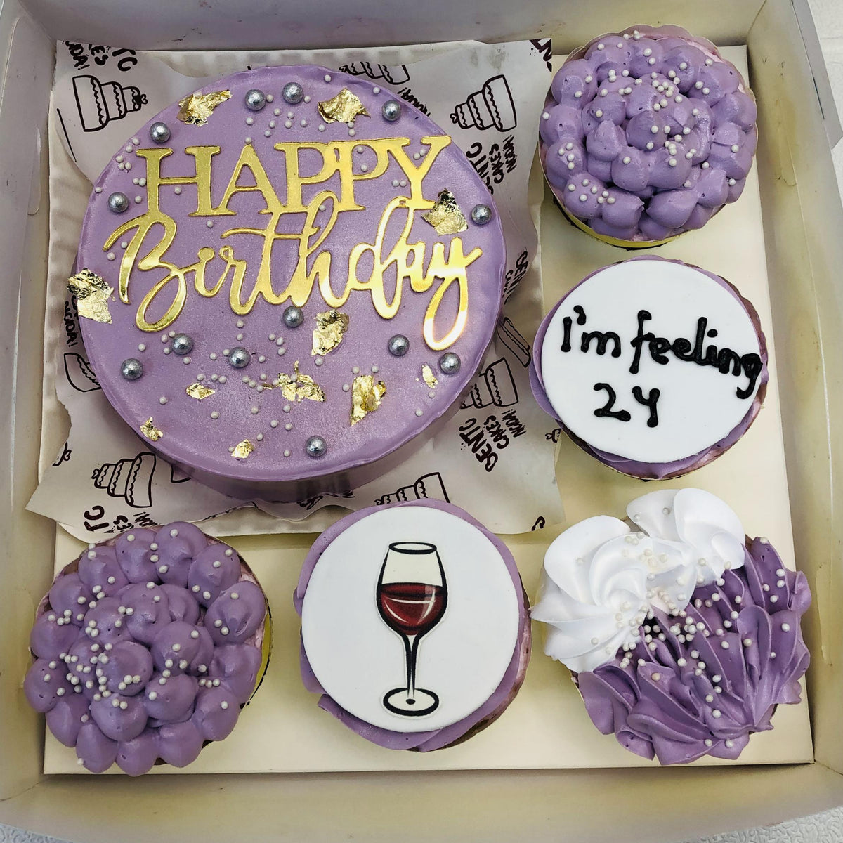 A set of purple-themed bento cakes and cupcakes arranged in a box. The centerpiece is a round cake with smooth purple frosting, decorated with gold leaf, silver pearls, and the words "HAPPY Birthday" written in gold. Surrounding the cake are five cupcakes with unique designs, including text, sprinkles, and a wine glass illustration.