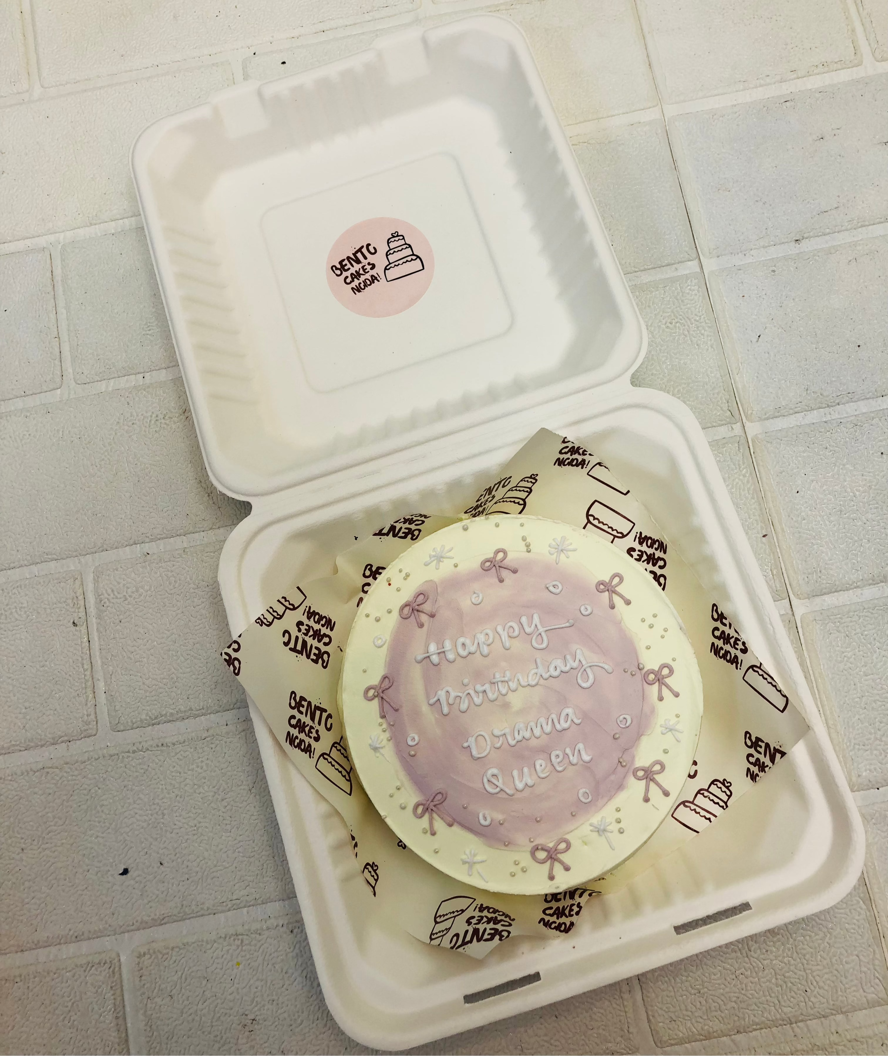 An offwhite bento cake having purple bows drawing all over the cake with purple shady design in center.