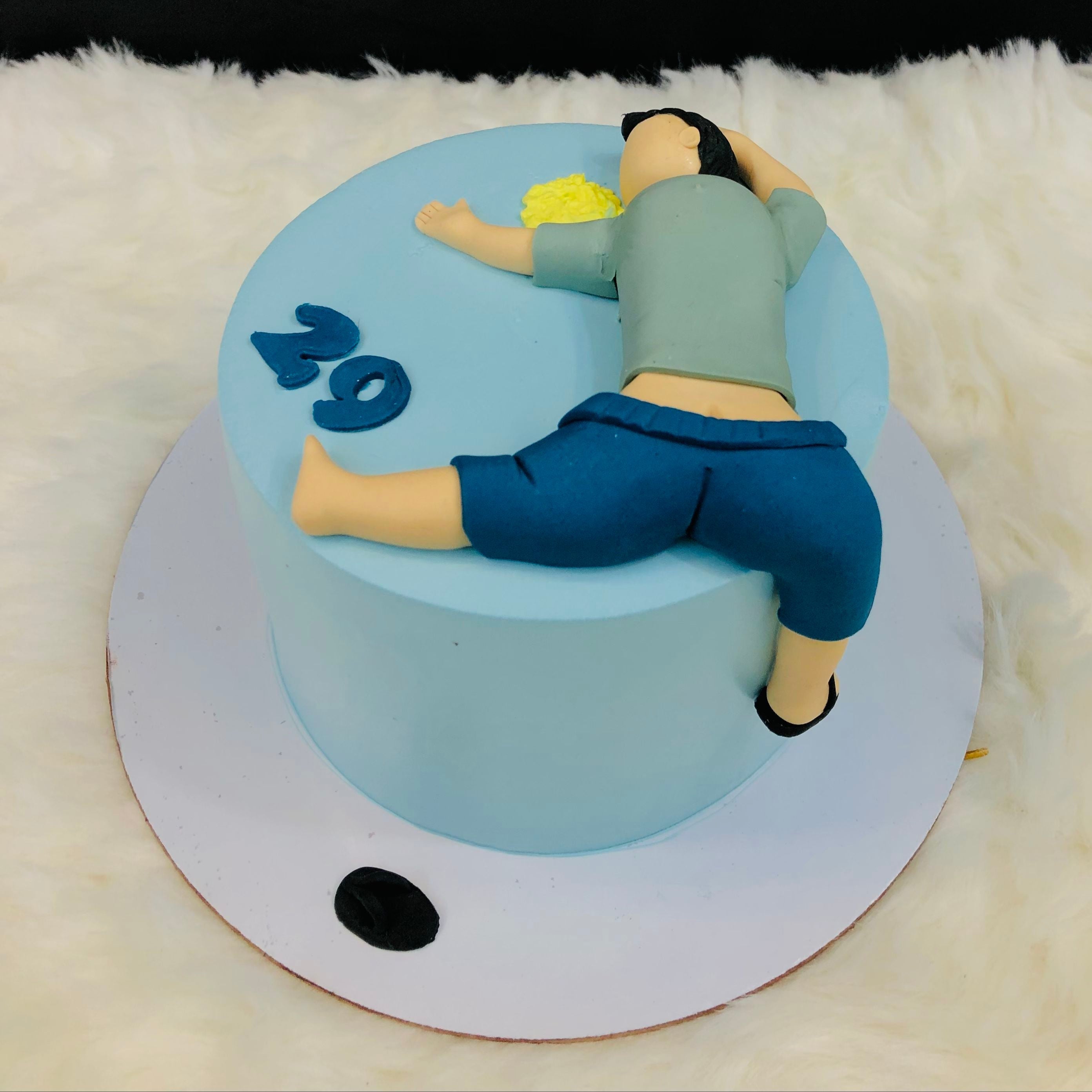 A round blue cake with a fondant figure of a person lying face down on top. The figure is wearing a green shirt and blue shorts, with one leg hanging off the side of the cake. The number "29" is also made of fondant and placed on the cake.