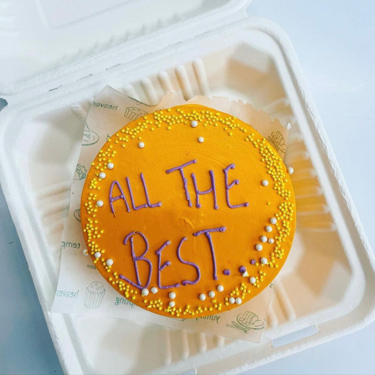 A round, orange cake with the words "ALL THE BEST" written in purple icing, decorated with yellow and white sprinkles, placed inside a bento box.