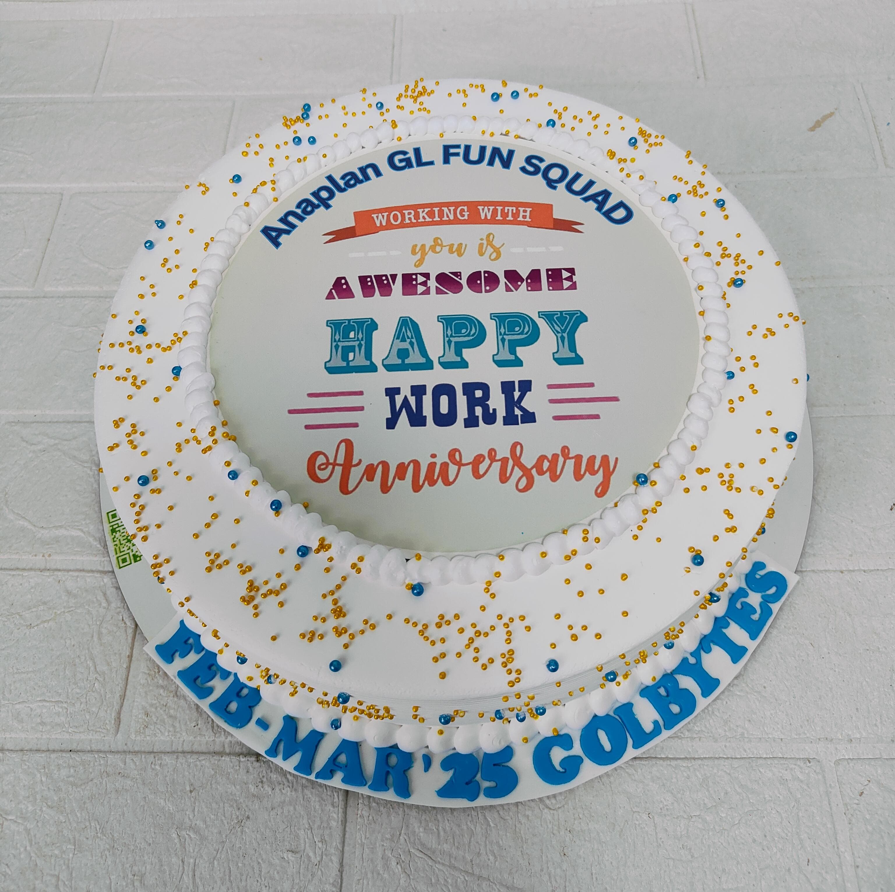 A round cake with white icing, adorned with gold and blue sprinkles. The cake features a colorful celebratory message for the Anaplan GL Fun Squad’s work anniversary, along with the text 'Feb-Mar '25 Golbytes' on the side.