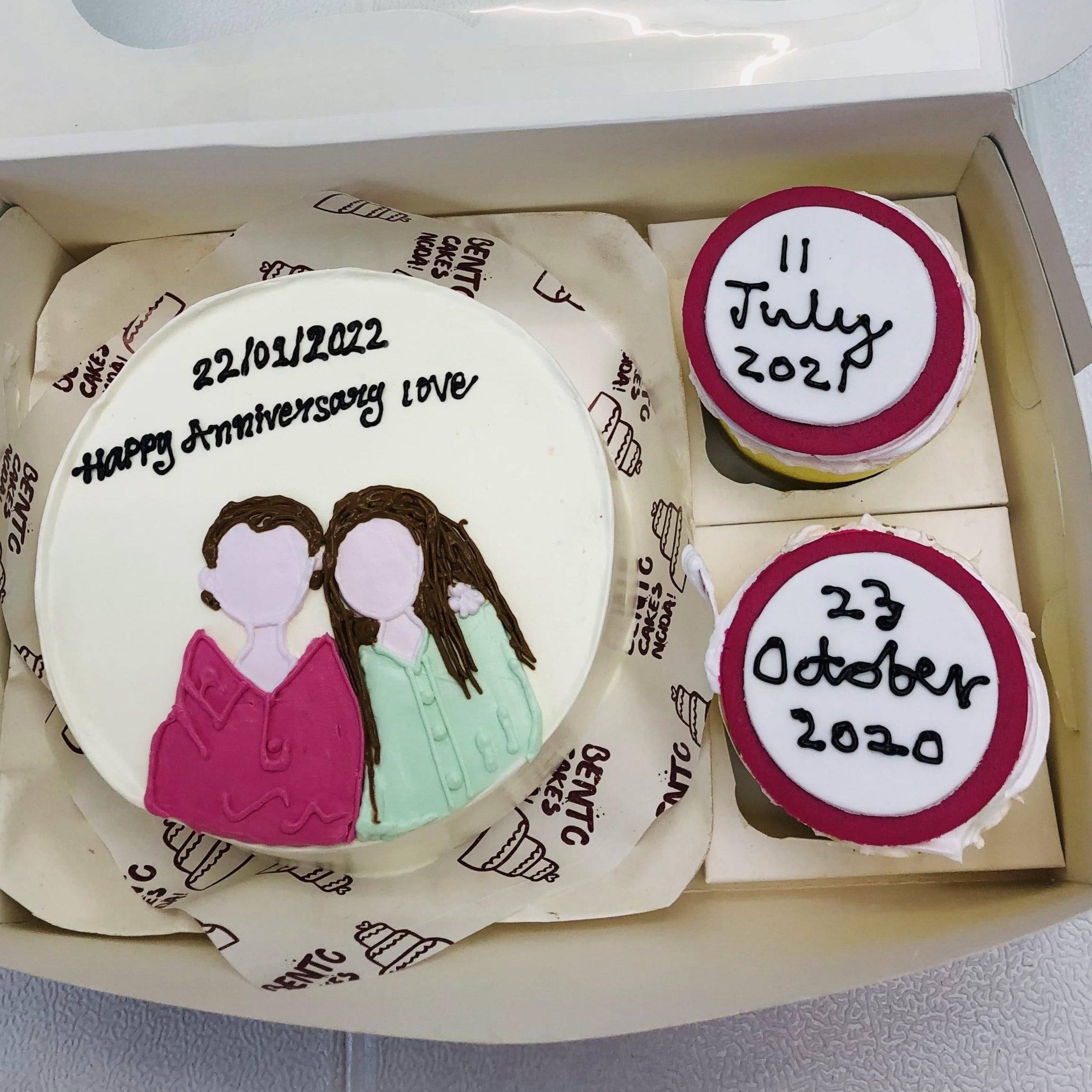 A mini bento cake set for anniversaries, featuring a main cake with an illustration of two faceless people and the text "22/01/2022 Happy Anniversary Love," accompanied by two smaller cupcakes with dates "11 July 2021" and "23 October 2020," all presented in a decorated box.