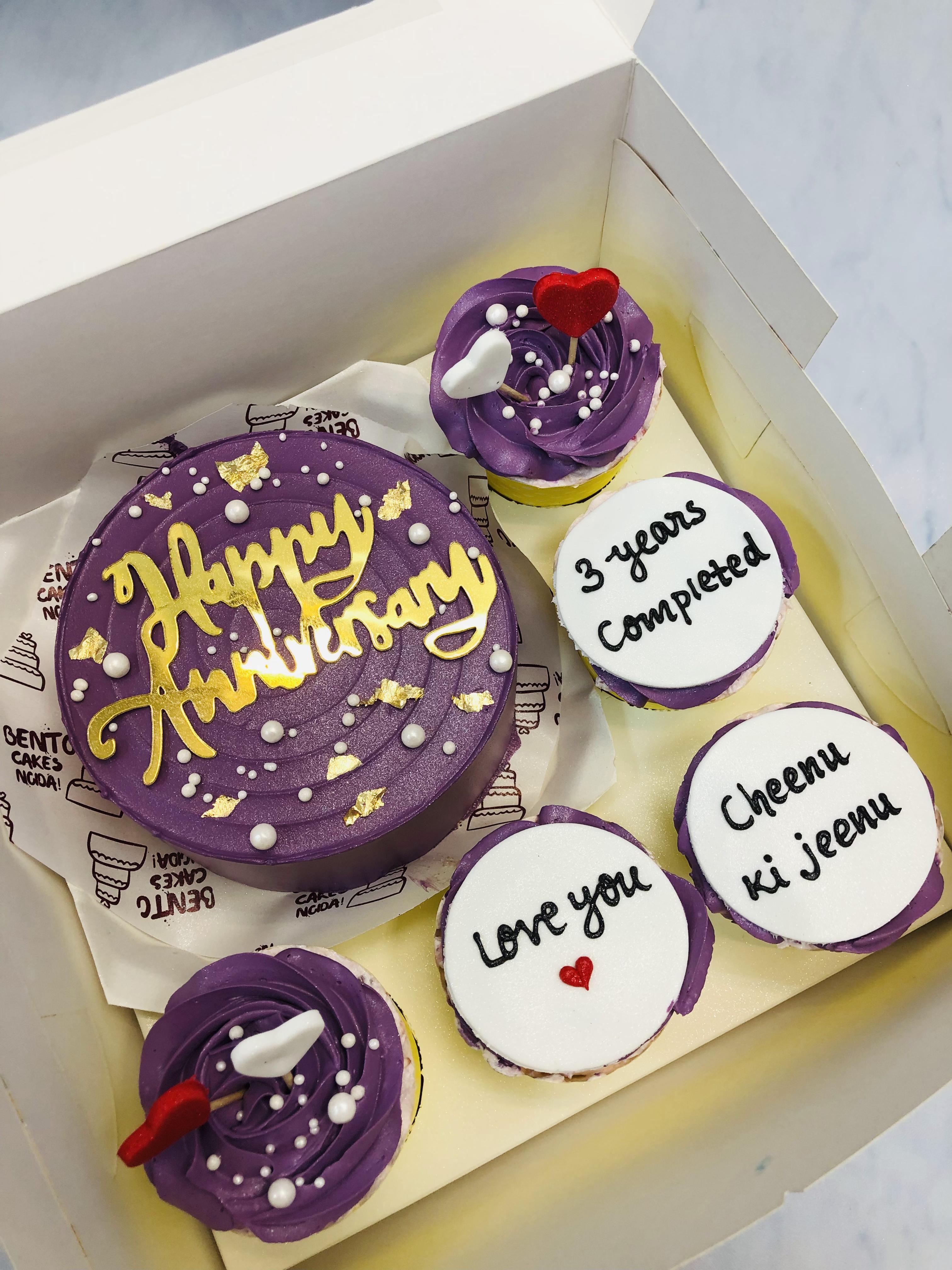 A purple-themed round anniversary cake with "Happy Anniversary" written in gold icing, surrounded by four cupcakes with messages like "3 years completed," "Love you," and "Cheenu ki jeenu." Two cupcakes feature purple frosting, white pearls, and red heart toppers.