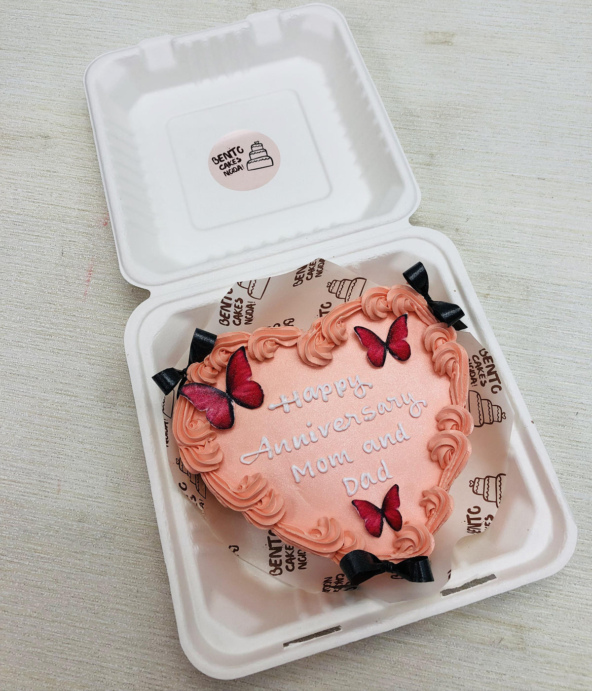 A shiny peach color heart shape bento cake with black bows and pink butterflies on it for mom dad anniversary.