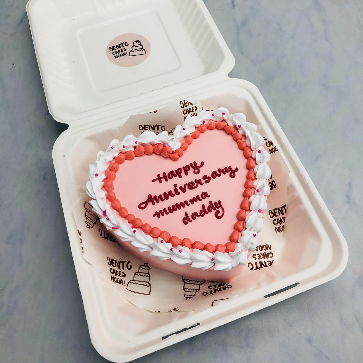 A light pink heart shape cake with happy anniversary text on it.