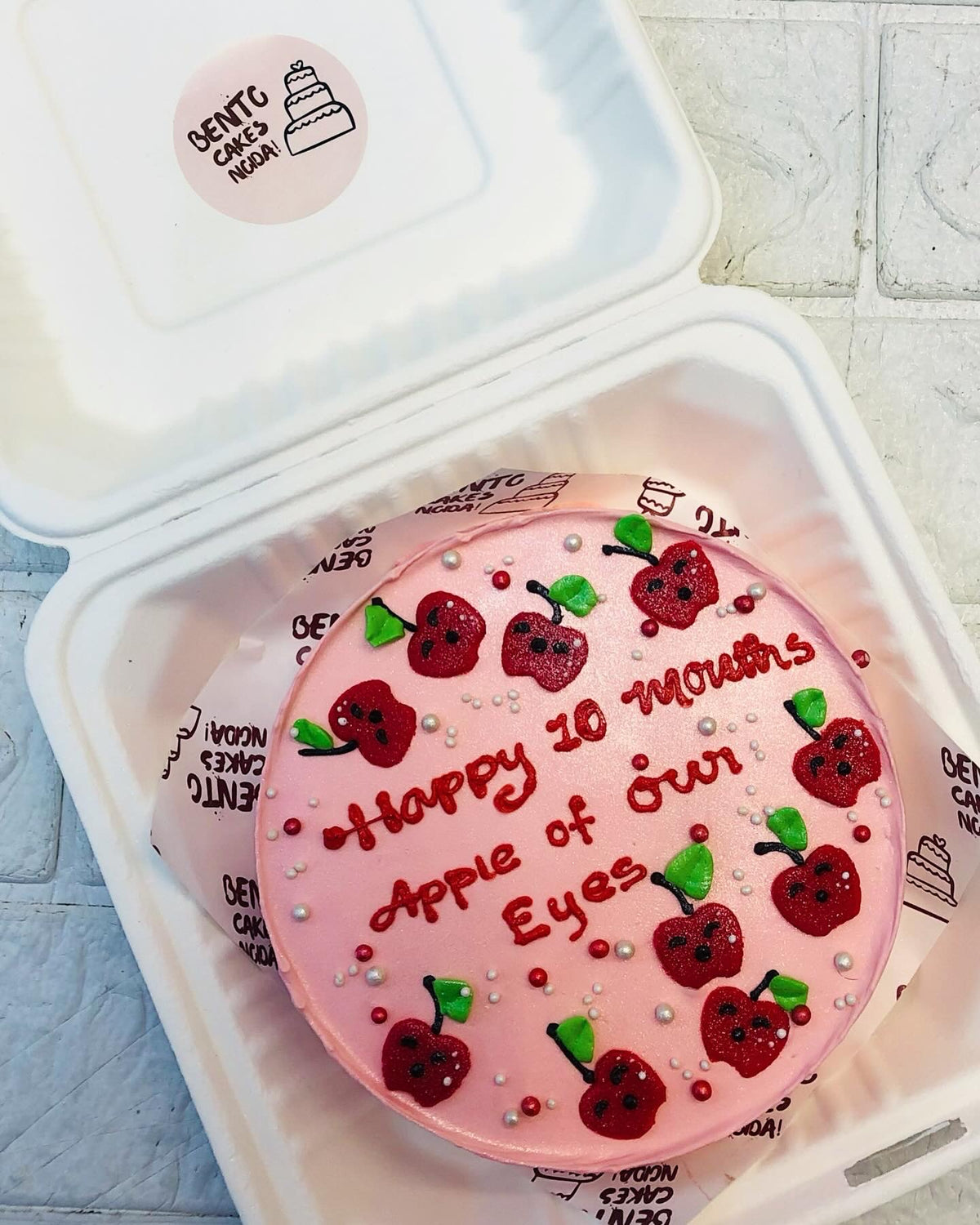 A light pink bento cake with cute little apples designs all over the cake and a loving text written on it.