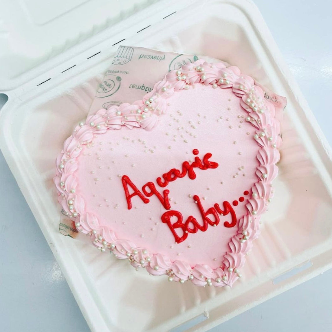 A pink heart-shaped cake decorated with the words "Aquarius Baby" written on top in elegant frosting.