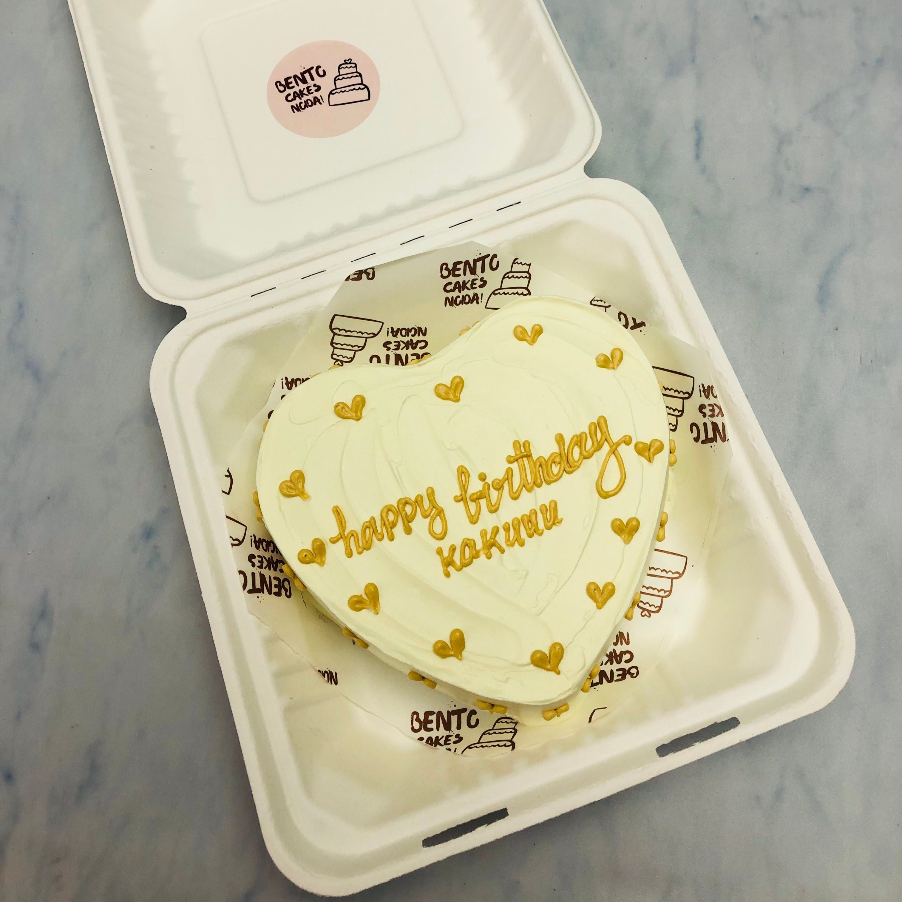 An attractive white heart shape bento cake with yellow hearts all ver it and a text "happy birthday kakuuu" with yellow icing on it.
