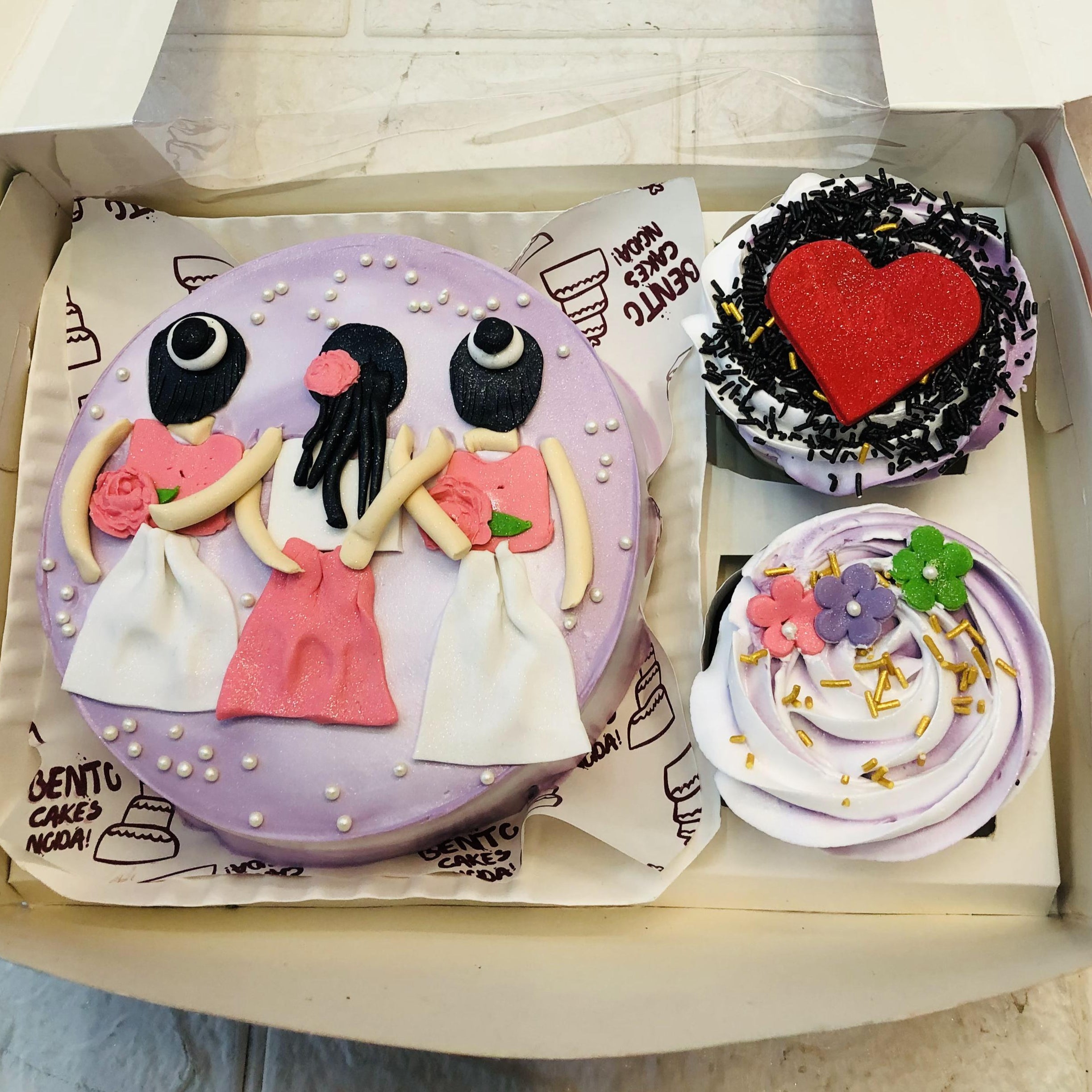 A "BFF Mini Bento Combo Cake" set featuring a main round cake decorated with three black-haired figures in pink and white dresses holding hands, symbolizing friendship. The cake is adorned with white pearl decorations. Two cupcakes accompany the cake: one with a red heart and black sprinkles, and the other with purple and white swirls, topped with colorful flowers and gold sprinkles.