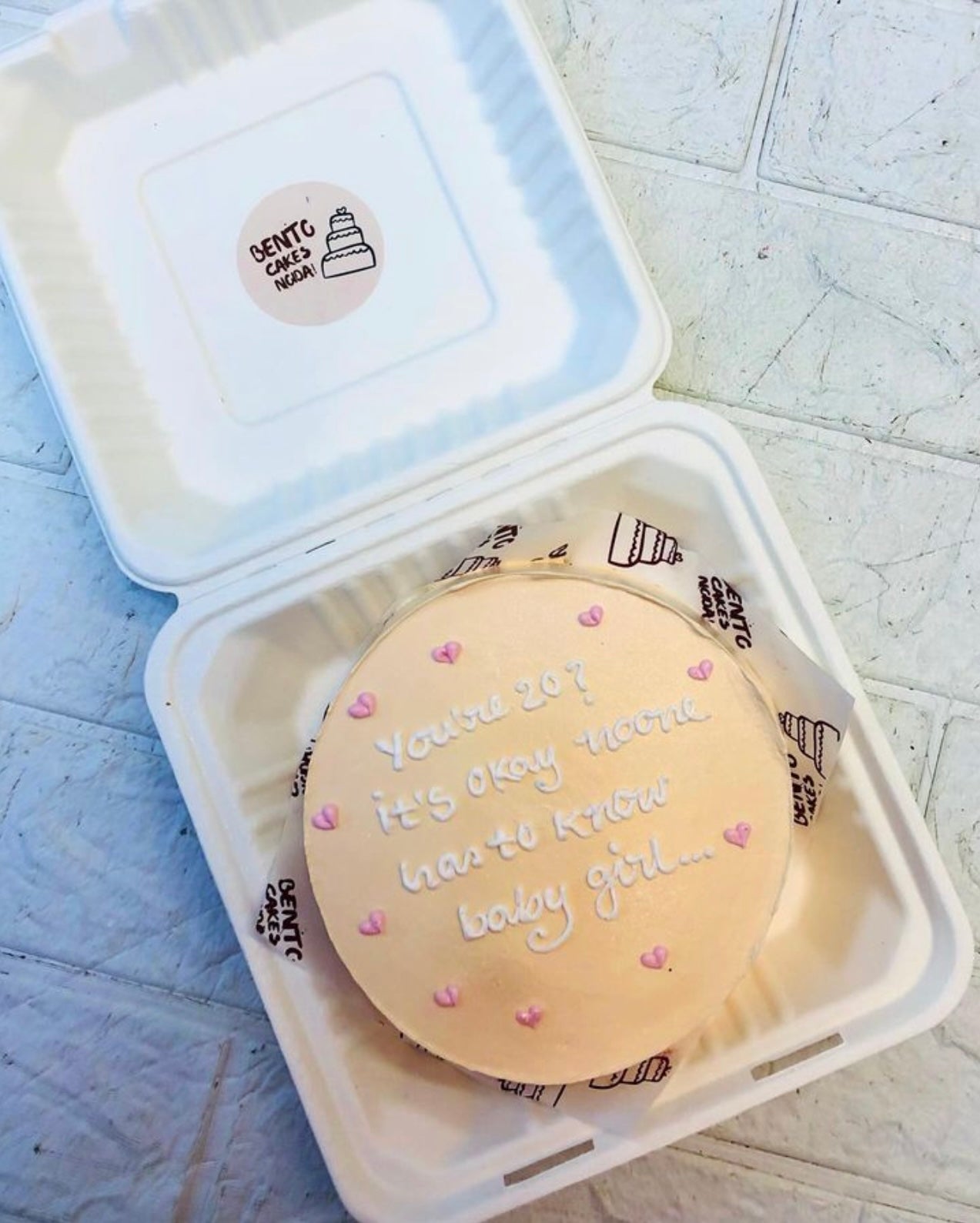 A peach color cake in a bento box decorated with light pink hearts design and a text written with white icing.