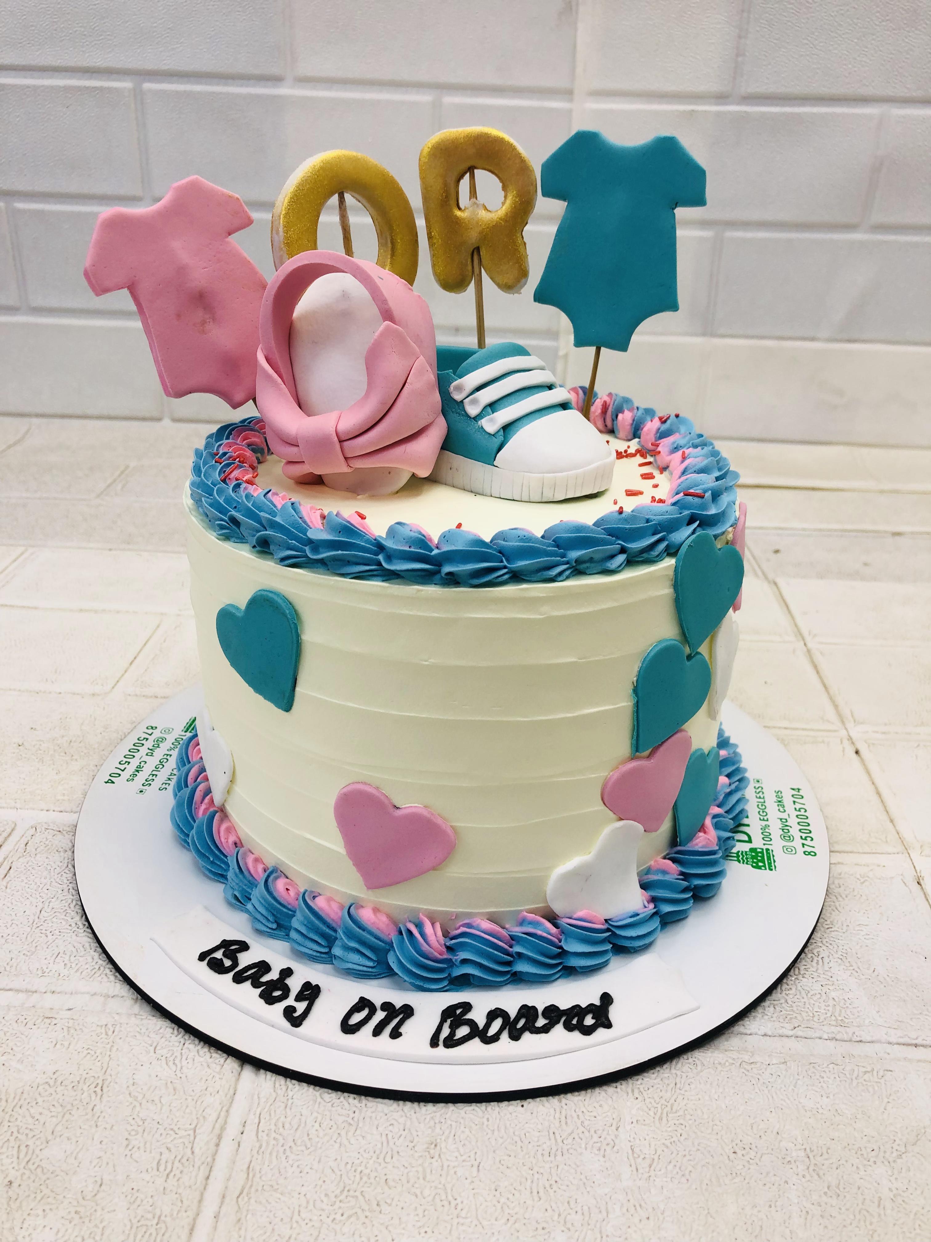 A beautifully decorated baby shower cake with a "Baby On Board" theme. The cake is adorned with fondant decorations including a pink baby onesie, a blue baby onesie, a pink baby shoe, a blue baby shoe, and golden letters spelling "OR". The cake is decorated with blue and pink hearts and swirls of blue and pink frosting.
