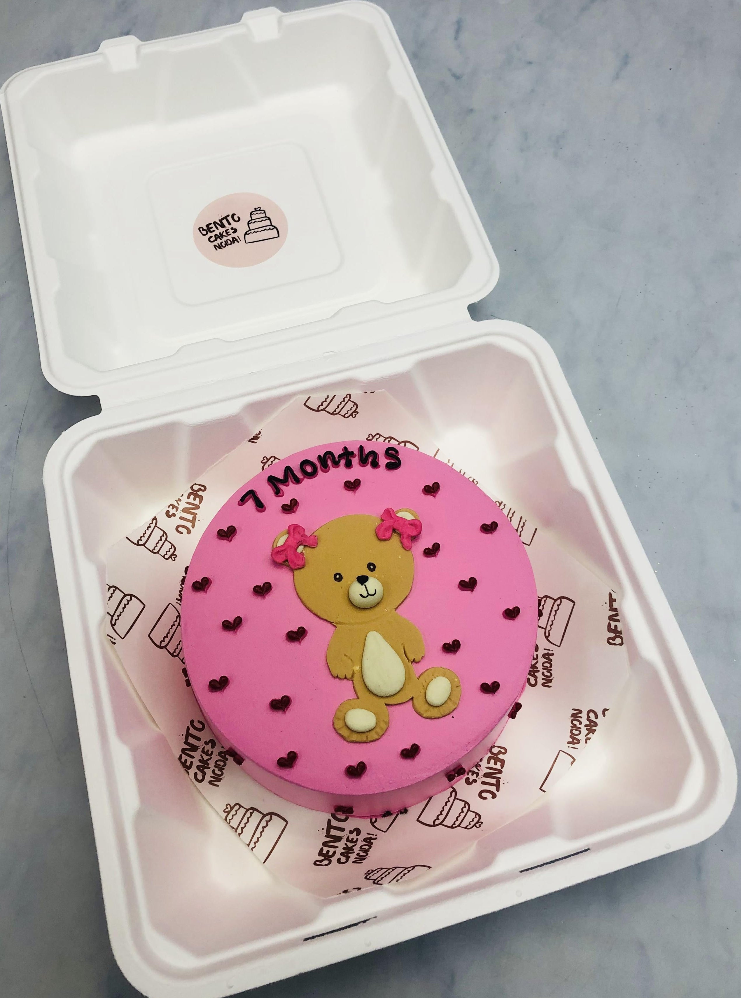 A light pink bento cake having a cute little teddy design and filled with red hearts for 7 months baby.