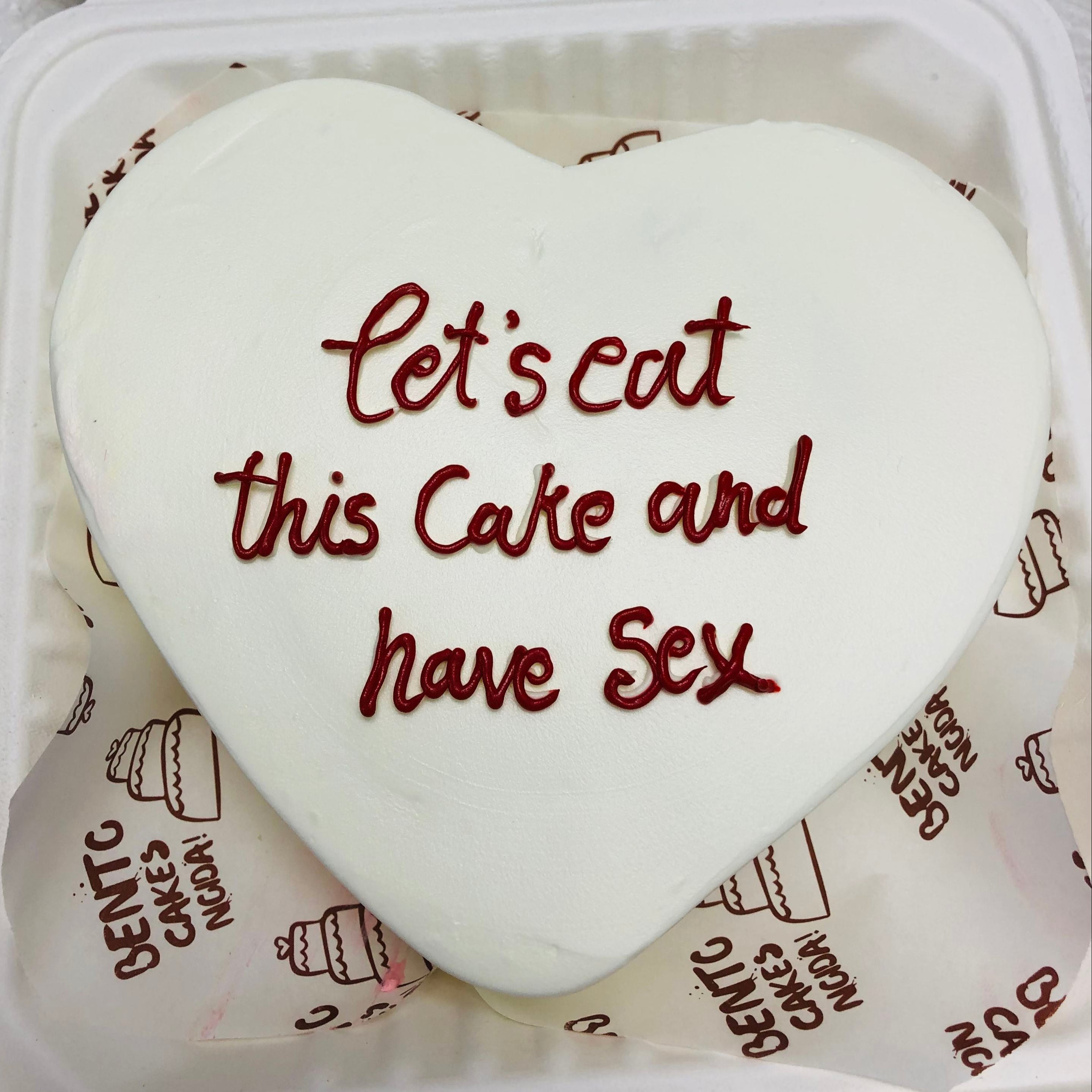 A heart-shaped cake adorned with the phrase "let's eat this cake and have sex" in decorative icing showing top view.

