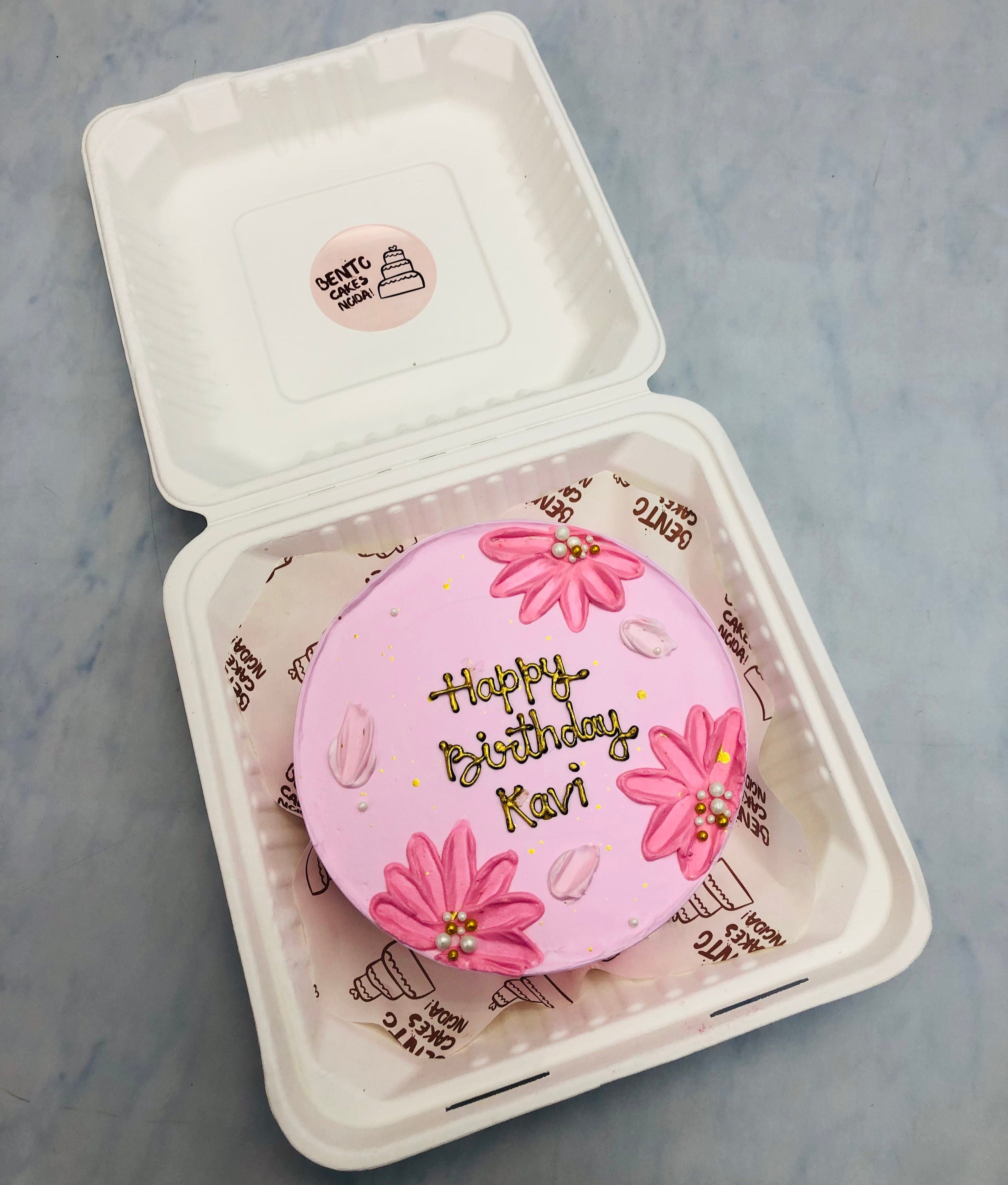 A pink birthday cake with floral decorations, placed inside a white bento cake box. The cake is decorated with pink flowers and petals, and has the message 'Happy Birthday Kari' written in gold icing. 